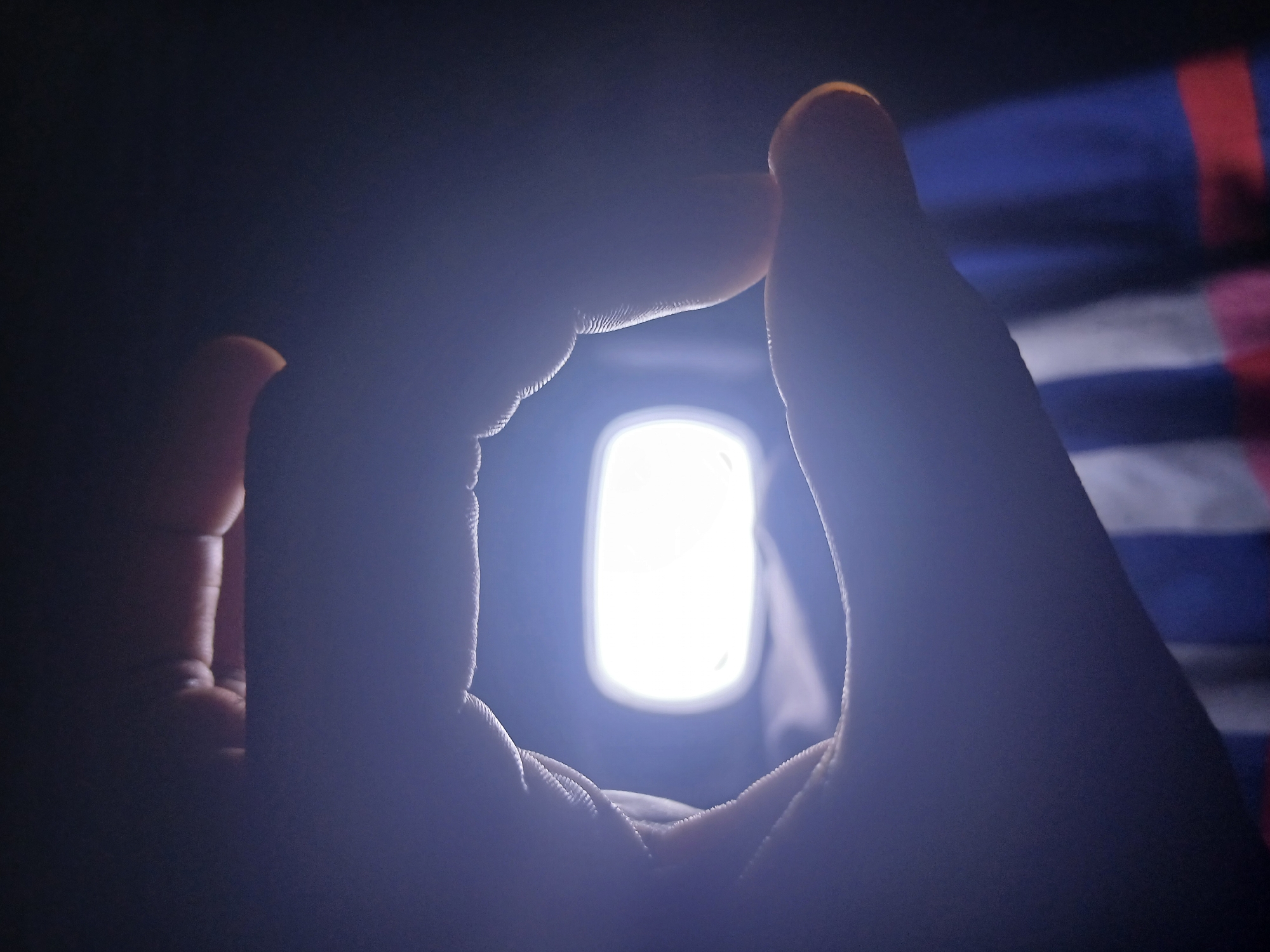 A person's hand is forming a square around a bright, rectangular light source. In the background, there are blurred stripes of blue, white, and red. The rest of the scene is in deep shadow. The location is not discernable from the image alone.
