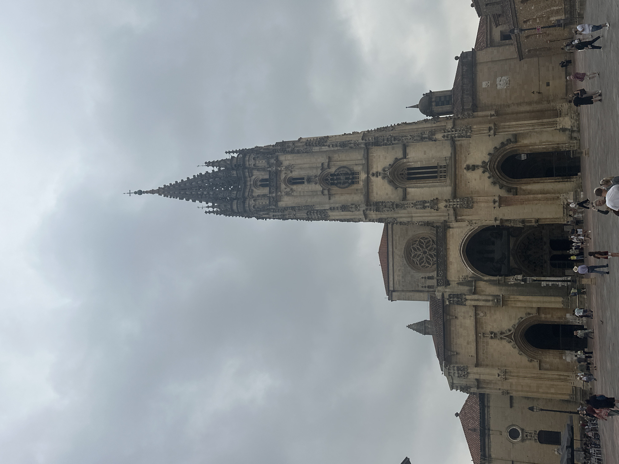 The image depicts the exterior of the Oviedo Cathedral in Oviedo, Spain, under a cloudy sky. The imposing Gothic structure features a tall, ornate tower with intricate stonework and a spire topped with a cross. The main facade of the cathedral is visible, with large arched doorways and detailed carvings. A rose window is situated above the central entrance. The cathedral is made of light-colored stone. In the foreground, a paved plaza is populated by a number of people. Some are walking, while others are gathered in small groups, appearing to be tourists or locals. There are outdoor tables and umbrellas visible to the left, suggesting a cafe or restaurant area. The overall atmosphere is that of a public square in front of a significant historical monument.