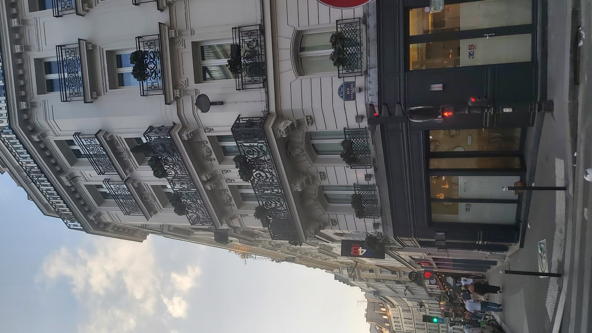 The image shows a street scene in Paris, France. A multi-story building with ornate balconies and windows dominates the right side of the frame. The building is light-colored and appears to be residential or mixed-use. On the ground floor, there is a dark storefront with large glass windows. A street sign for "9e Arr. RUE BERGERE" is visible on the side of the building.
To the left of the building, a street extends into the distance, lined with more buildings, some of which have illuminated signs. Pedestrians are walking on the sidewalk. A traffic light with a red pedestrian signal is visible in the foreground, and a green traffic light is further down the street. The sky is partly cloudy.