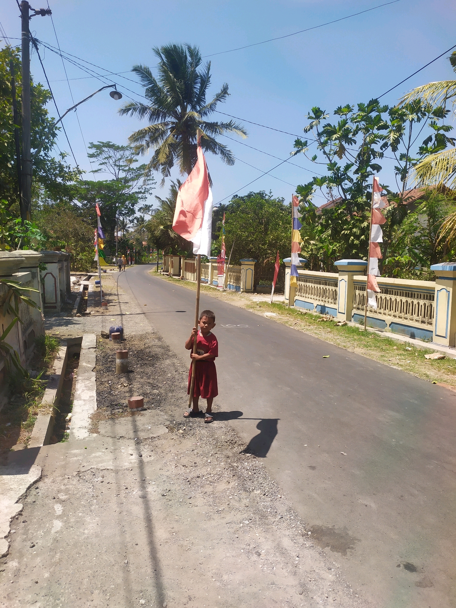 A young child, appearing to be of Southeast Asian descent, stands on the side of a paved road in the village of Desa Penggalang, Kecamatan Adipala, Indonesia. The child, wearing a red dress and sandals, holds a long wooden pole with an Indonesian flag attached to it. The flag is a horizontal bicolour of red over white. The road appears to be in a rural or semi-rural setting, with a concrete drainage ditch running alongside it on the left. A decorative fence, painted yellow and blue, lines the right side of the road, with several multi-colored pennant flags also displayed along it. Lush green trees and vegetation are visible in the background, along with houses and power lines. The sky is clear and blue, indicating a sunny day. The child's shadow is cast on the road.