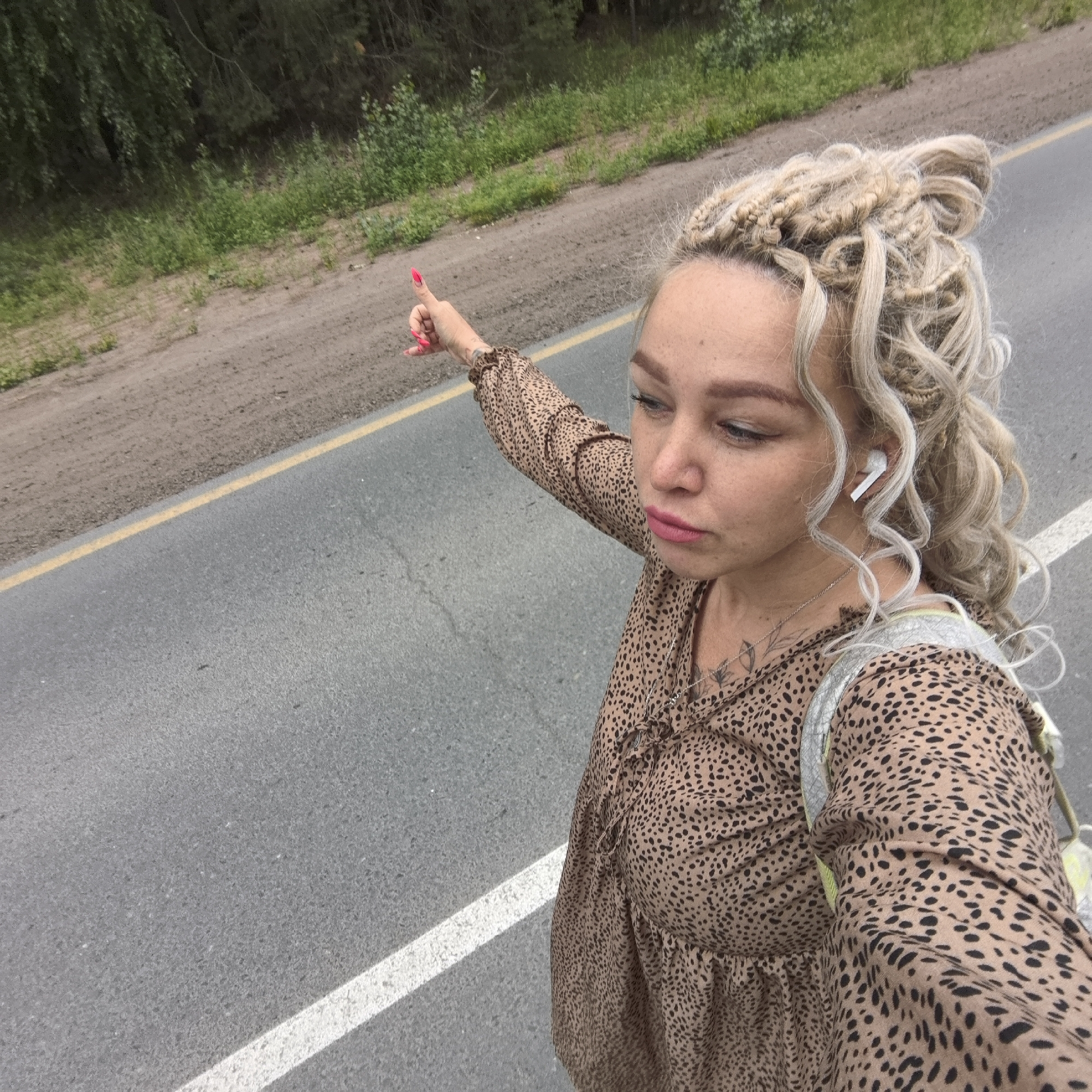 A woman with blonde braided hair and light skin, wearing a leopard-print dress, stands on the side of a road in Noviy, Russia. She is wearing white wireless earbuds and has a backpack on her shoulder. Her left arm is raised, with her thumb pointed upwards, suggesting she might be hitchhiking. The road has a yellow and white line marking its edge. The background consists of a dirt shoulder and lush green vegetation, with trees in the distance. The image appears to be a selfie taken from a high angle.