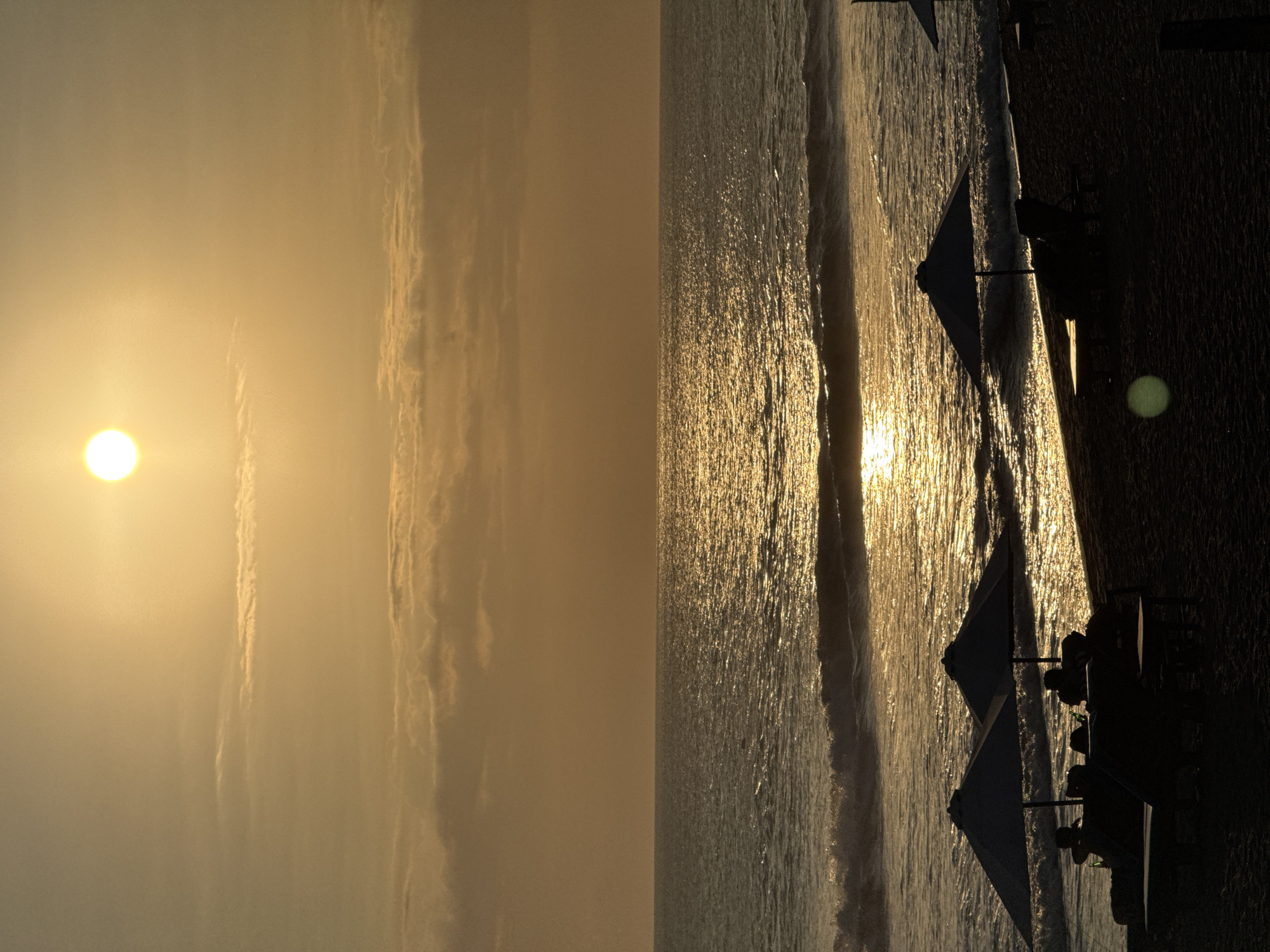 The image is divided into two scenes. The left scene depicts a golden sky with the sun visible, likely at dawn or dusk. Wispy clouds stretch vertically across the scene. The right scene shows a beach with waves reflecting the sunlight, creating a shimmering effect on the water's surface. Umbrellas are set up along the beach, and people are sitting beneath them. The scene is dark, and people and objects are displayed as dark silhouettes. The location appears to be North Kuta, Indonesia, based on the beach and tropical environment.
