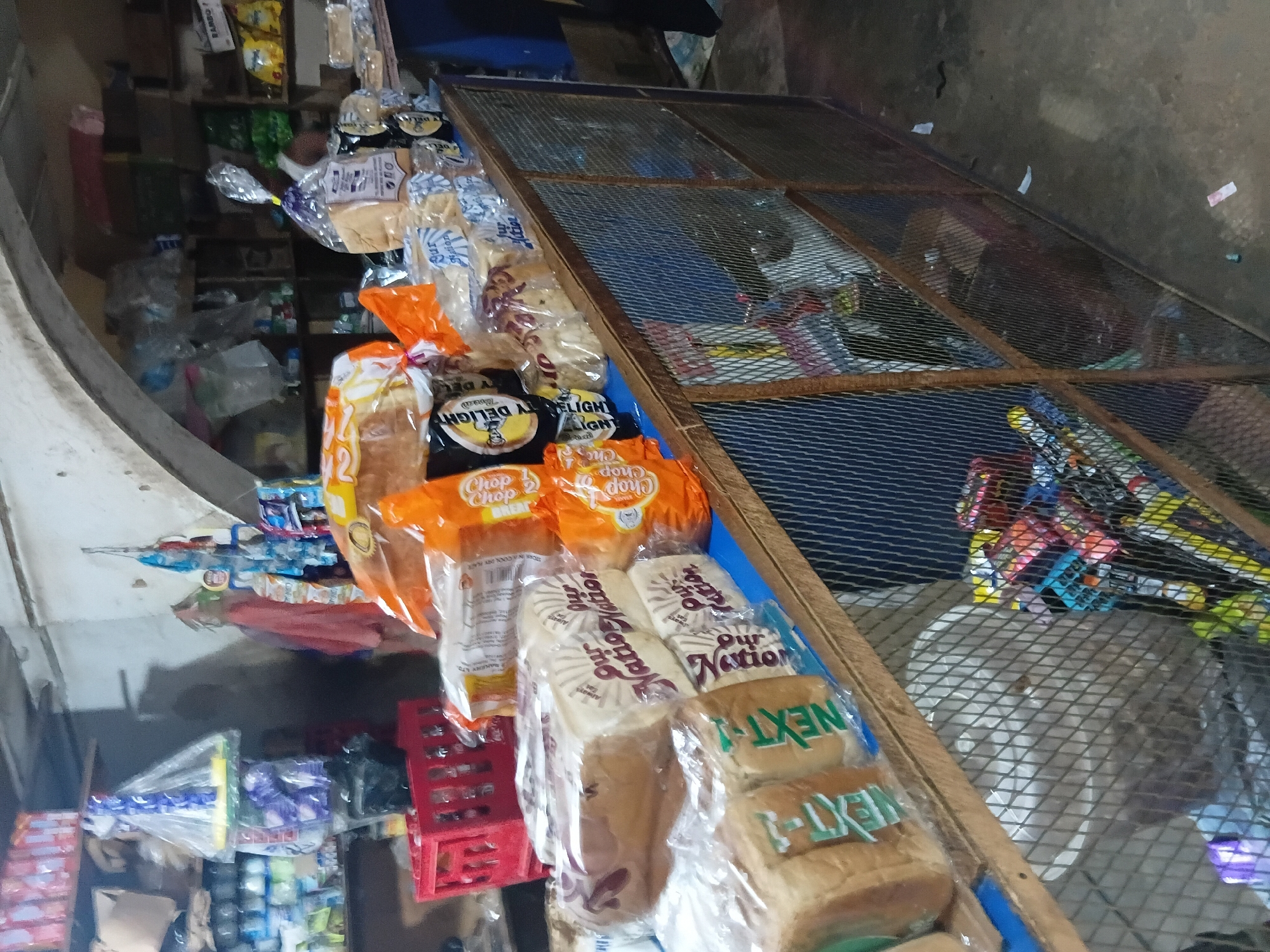 The image shows a small retail store, likely a provisions shop, in Jos, Nigeria. The shop is cluttered with various food items, primarily loaves of bread packaged in plastic. These loaves are stacked on shelves and a counter. Some of the bread packaging displays brand names like "Our Nation" and "NEXT-1". Other items visible include red plastic crates, bags of snacks or confectionery, and what appear to be drinks in plastic bottles on shelves in the background. The store has a rustic appearance, with wooden shelves and a counter, and a wire mesh fence in the foreground. The lighting is dim, suggesting an indoor setting with artificial illumination or natural light from an opening. The overall impression is of a typical small business operating in an urban or semi-urban environment in Nigeria, selling everyday consumer goods.