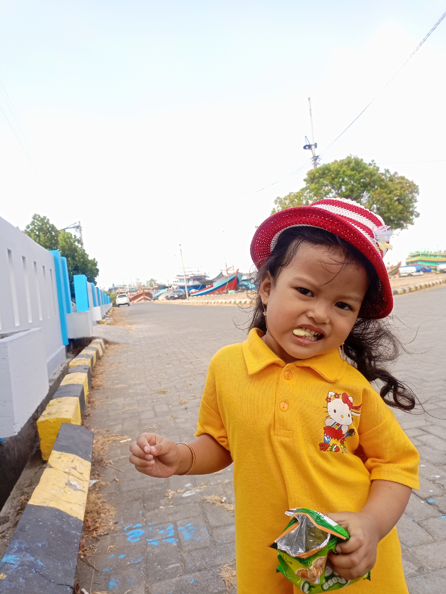 A young girl with dark hair and a red and white striped hat stands on a paved walkway in Juwana, Indonesia. She is wearing a yellow polo shirt with a Hello Kitty graphic. Her mouth is open, and she is holding a partially eaten bag of chips. In the background, there is a street with parked cars and several boats docked in the water, suggesting a port or harbor area. The walkway is bordered by a low wall painted in alternating grey, yellow, and black stripes. The sky is overcast.