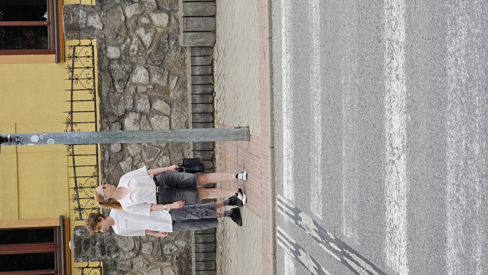 The image depicts two individuals standing on a sidewalk next to a pedestrian crossing. A young male, likely a teenager, stands to the left wearing a white t-shirt, grey cargo shorts, and black sneakers. He has on bright yellow sunglasses. To his right, a woman stands wearing a textured white collared shirt, dark grey denim shorts, and black and white sneakers. She has a black belt with a gold buckle and is holding a black handbag. Both individuals appear to be facing towards the street, possibly waiting to cross. Behind them, a stone wall topped with a decorative black metal fence runs along the property line. A yellow building with a dark-framed window is visible above the fence. A utility pole with stickers on it stands to the right of the individuals. The ground consists of a paved sidewalk and a grey asphalt road with white pedestrian crossing markings. The scene appears to be a typical urban streetscape, consistent with a location in the city of Prešov, Slovakia, given the architectural style and the presence of a pedestrian crossing.