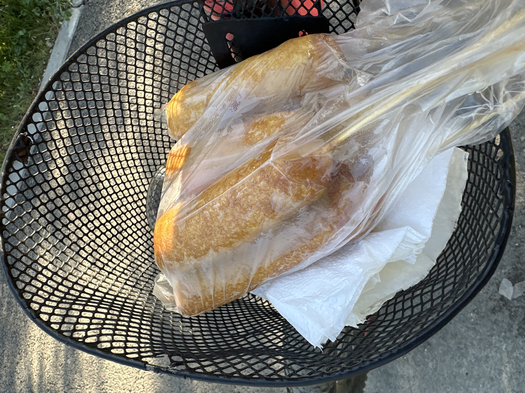 Captured from a high-angle perspective, the photograph presents a black wire mesh waste bin located in the city of Unknown, Unknown. Inside the bin, a plastic bag encases a pile of what appears to be deep-fried food, possibly corn dogs, arranged on top of some crumpled white paper napkins. The bin itself is situated outdoors, resting on a concrete surface near a patch of green grass. The light suggests it is daytime.