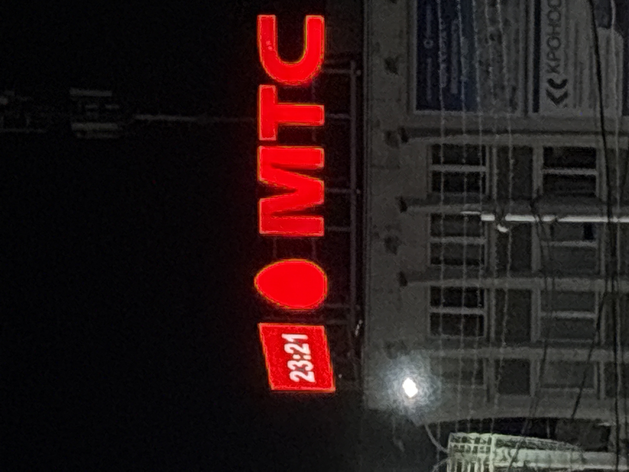An illuminated sign displays the time "23:21" and the letters "MTC" in bright red neon. The sign is mounted on a building at night. A transmission tower is visible in the background. The building appears to be a commercial structure with multiple windows. The scene is located in the city of Vitebsk, Belarus.