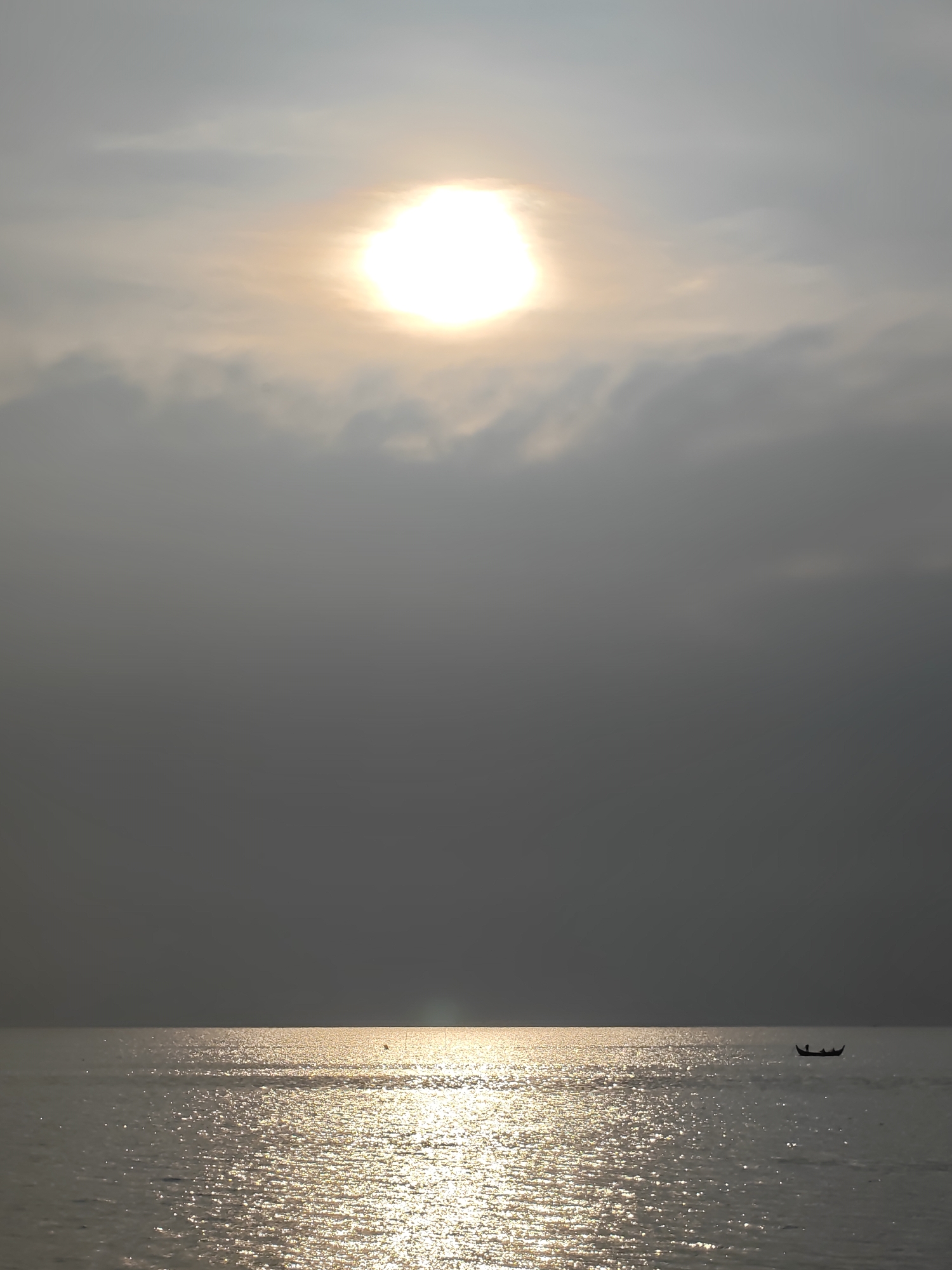 The image depicts a serene maritime scene, likely at sunrise or sunset, characterized by a glowing sun over a body of water. The sky is largely covered by grey clouds, through which the intensely bright sun shines, creating a vibrant white and golden halo. Below the horizon, the water's surface reflects the sun's light, forming a shimmering path of gold and white ripples that extends towards the viewer. A small, dark silhouette of a traditional boat, possibly a fishing boat, floats peacefully on the right side of the water, near the horizon. Within the boat, two indistinct dark figures are visible. The overall atmosphere is tranquil and calm, with the cloudy sky and shimmering water dominating the frame. The scene is set in Lhokseumawe, Indonesia. No text is visible in the image.