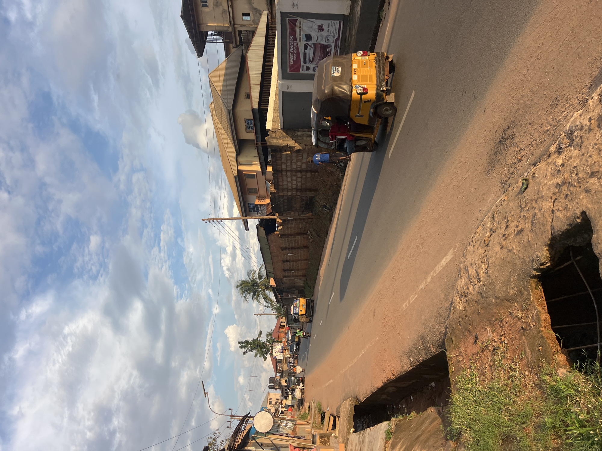 The image depicts a street scene in Nkpor, Nigeria, likely during daylight hours given the presence of shadows and the brightness of the sky. The sky is filled with scattered white clouds against a blue background, suggesting a partly cloudy day.
The scene is dominated by a paved road running diagonally through the frame. A yellow motorized tricycle, commonly known as an "Okada" or "Keke Napep" in Nigeria, is the most prominent object on the road, moving away from the viewer. Two people are visible inside the tricycle. A person in a blue shirt is riding on a motorcycle behind the tricycle.
Buildings line the side of the road. Some appear to be residential or commercial, with corrugated metal roofs and brick walls. A prominent billboard with text is attached to one of the buildings. The text visible on the billboard is "SWEET FINGERS FOOD CENTRE".
The road is bordered by a raised, somewhat dilapidated concrete structure on the right side, with a drainage ditch below it. This structure appears eroded and has patches of grass growing on it. The overall atmosphere suggests a busy urban or semi-urban environment with active street life. The angle of the photograph, taken from a slightly elevated position and rotated, gives a unique perspective on the street.
