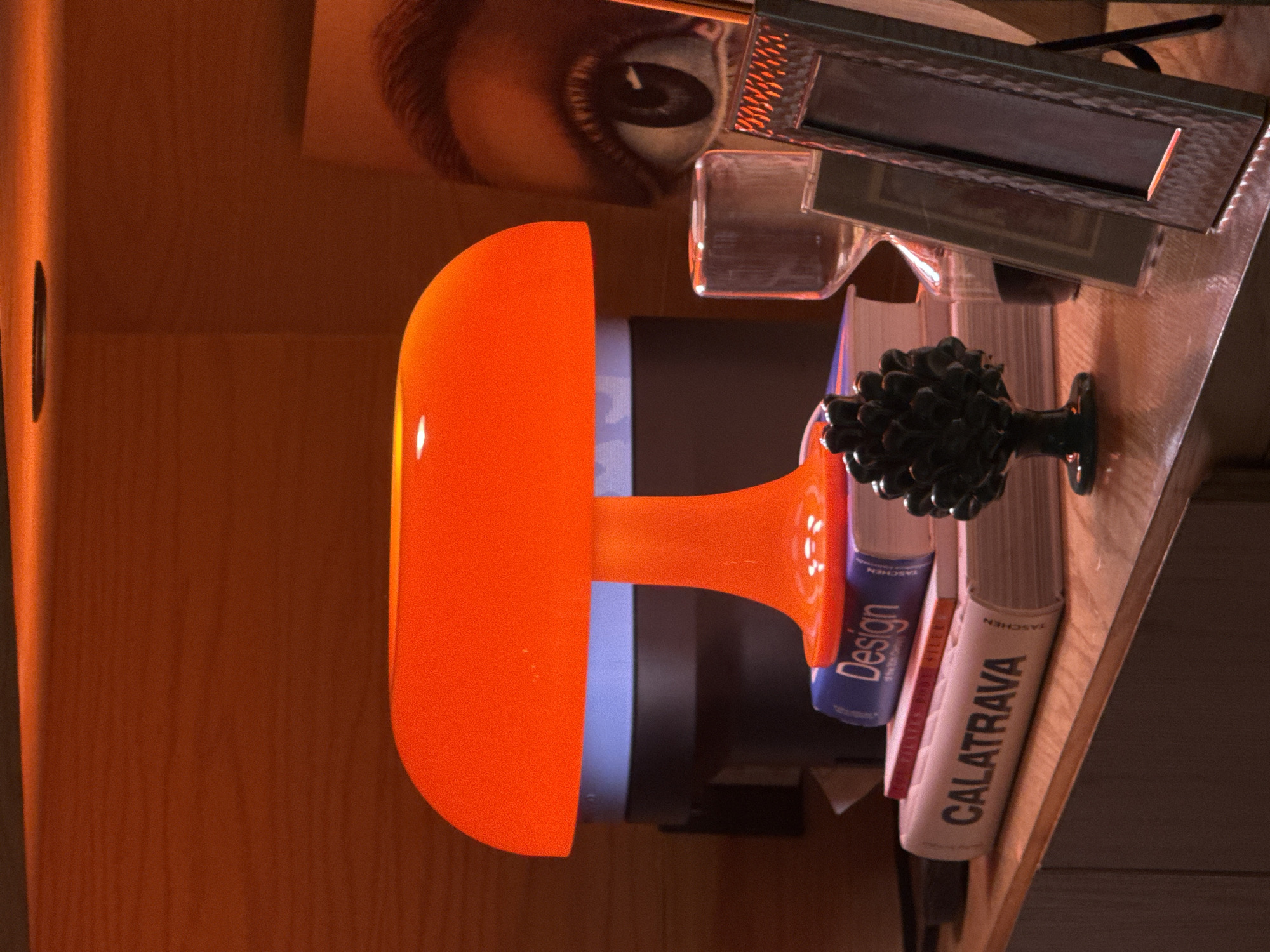 The image depicts a collection of objects arranged on what appears to be a wooden surface. Visible items include an orange lamp with a distinct rounded shape, books with visible titles such as "Design" and "CALATRAVA", a decorative object resembling a dark, ornate knob or handle, a pair of eyeglasses, and a partial image of an eye. The environment is dimly lit, contributing to the warm tones in the scene. There is no information about the location in the city of Puebla, Mexico.