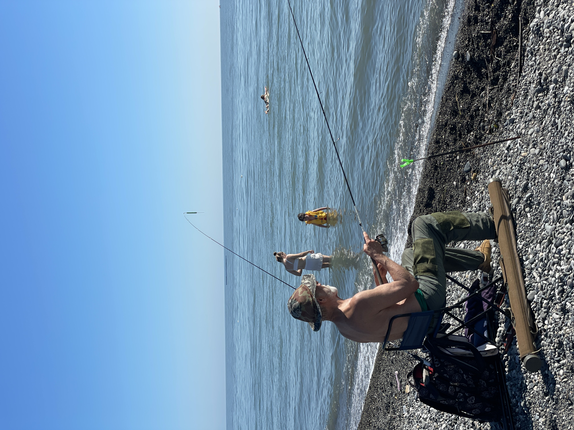 This is a clear, outdoor photograph taken on a sunny day at a pebble beach next to a large body of water in Unknown, Unknown. The sky is uniformly clear and bright blue.
In the foreground, an older male with a white beard and a camouflage bucket hat is seated on a dark blue folding chair, facing the water with his back mostly to the viewer. He is shirtless and wearing long green pants with dark patches on the knees, and light brown shoes. He is actively holding and operating a fishing rod with a reel. To his left, a dark-colored backpack with a floral pattern rests on the pebbles, next to what appears to be another fishing rod. A brown, cylindrical fishing rod case lies horizontally on the pebbles in front of his chair. To his right, closer to the water's edge, a dark-colored fishing rod holder with a bright green tip is staked into the pebbles.
Several individuals are in the water. In the mid-ground, a young adult female in a white two-piece swimsuit stands waist-deep, facing slightly away from the camera. To her right, a young child wearing a yellow life vest or flotation device stands chest-deep in the water, looking towards the shore. Further out in the deeper water, a person lies prone on a light-colored inflatable float.
The water is calm with small waves gently lapping onto the pebble shore. In the far left background, a dark, elongated object floats horizontally on the water's surface. A faint, dark shape, possibly a boat, is visible on the horizon in the far right background.