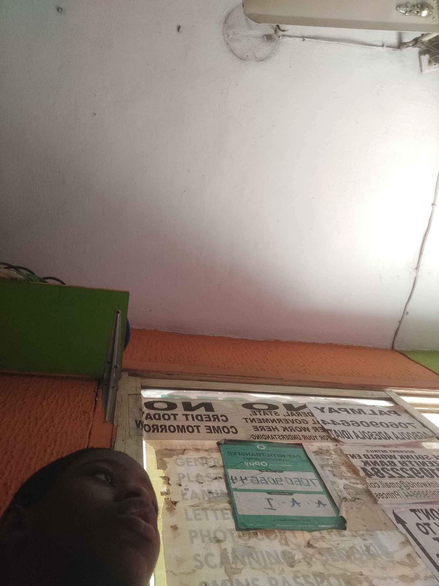 The image shows the lower half of a person's face in the lower left corner of the frame. Adjacent to the person is a window frame covered in advertisements and announcements. The ads and announcements include words like "CREDIT," "TRANSFER," "PAY WITH TRANSFER," "GENER," "LETTER," "PHOTO," "SCANNING," "PASSPORT," and what appears to be an address. The color scheme involves white, orange, and green. A green shelf or cupboard is visible above the window. The lighting is inconsistent, with some areas appearing bright while others are dim. The image appears to be taken indoors, potentially in a business or commercial space. The image's location is identified as Shangisha, Nigeria.