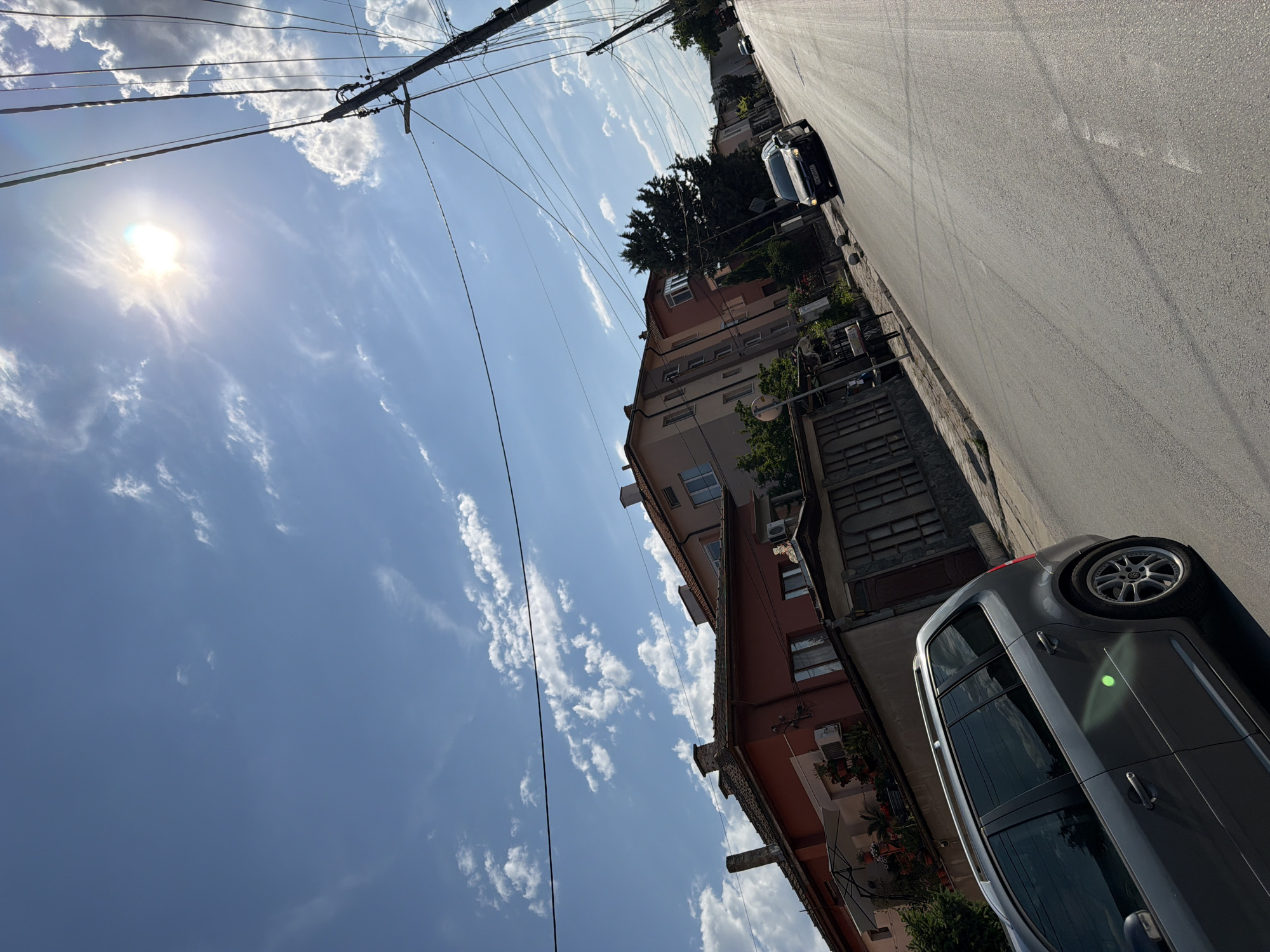 The image depicts a street scene in the city of Plovdiv, Bulgaria, under a bright, sunny sky with scattered clouds. The perspective is from a low angle, looking up and down a paved road lined with residential buildings and trees.
In the foreground, on the left side of the street, a dark grey station wagon is parked. The car has tinted windows and roof rails. Further down the street, on the right side, a black car is driving away from the viewer. There are utility poles with numerous wires running across the upper portion of the image.
The buildings on either side of the street appear to be multi-story residential structures, with some having red tile roofs. There are visible windows, balconies, and some greenery in front of the houses. The sunlight creates glare on the road and casts shadows. The overall atmosphere is that of a typical sunny day in a residential area.