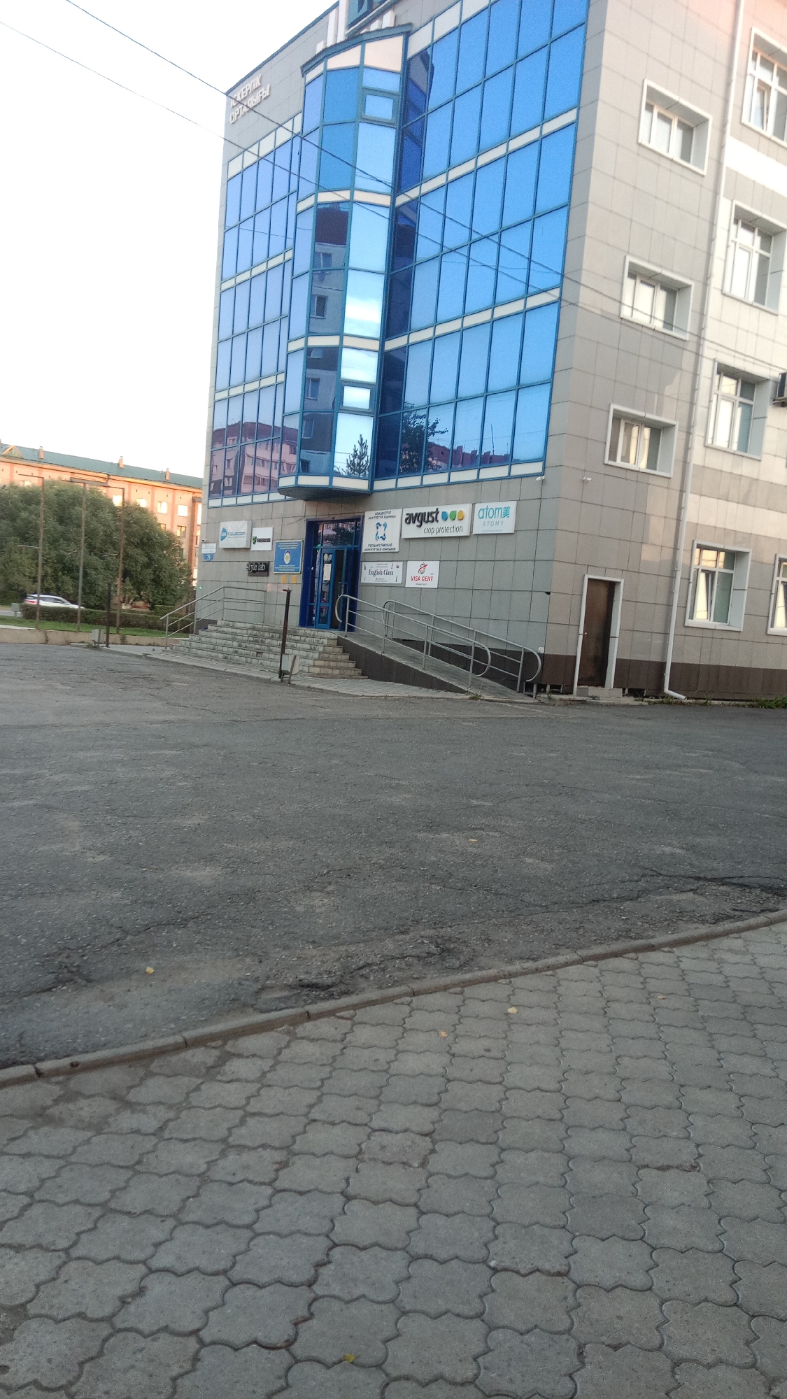 The image shows the exterior of a modern, multi-story commercial building in the city of Petropavl, Kazakhstan. The building features a facade of light-colored grey panels and large, blue-tinted glass windows. A prominent sign on the building reads "AVGVST" and "ATOM," suggesting these are business names. The entrance to the building is visible, with a set of stairs and a wheelchair-accessible ramp leading up to double glass doors. Several other signs are also visible near the entrance, including one that says "English Class" and another indicating "VISA CENT." To the left of the building, another structure with an orange facade and trees can be seen. The ground in front of the building is paved with asphalt in the foreground, transitioning to patterned grey pavers. The scene appears to be a public space outside of a business district, likely during daylight hours given the lighting conditions.