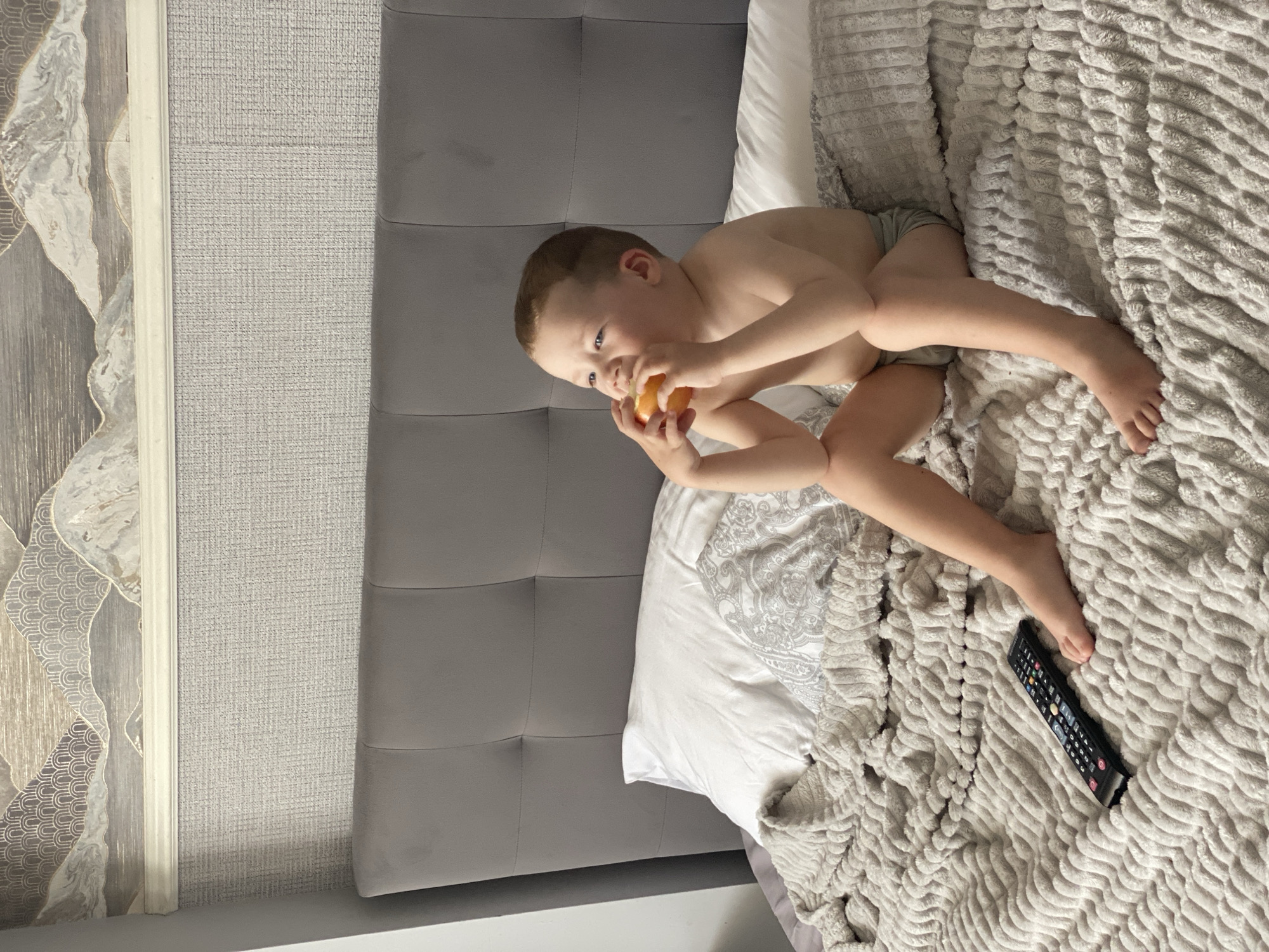 The image shows a young boy lying on a bed, eating an apple. He is wearing a diaper and is partially covered by a plush gray blanket. A TV remote control is visible on the blanket near his feet. The bed is in a room with a textured gray headboard. To the left of the bed is a wall with decorative wallpaper featuring abstract mountain-like patterns in shades of gray and beige. The room appears to be well-lit, suggesting it is daytime. The boy's expression is one of quiet contentment as he enjoys his snack. The overall scene is domestic and relaxed, likely taking place in a home. There is no visible text in the image.