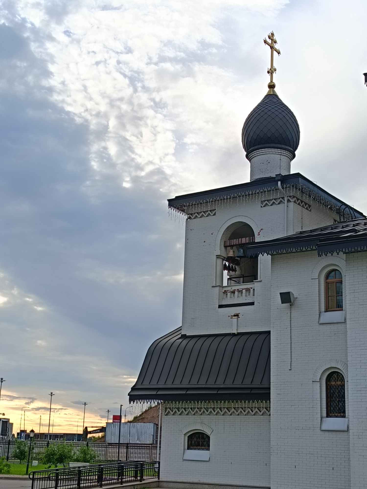 The image shows a white brick church with a black domed roof and a gold cross. The church has a bell tower with bells visible. The sky is cloudy with rays of sunlight breaking through. In the background, there are industrial buildings, streetlights, and a fence. The setting appears to be an urban area, likely in Saint Petersburg, Russia, given the architectural style and the presence of a construction vehicle. There are no people or animals visible in the image. The overall scene depicts a religious building within a city environment.