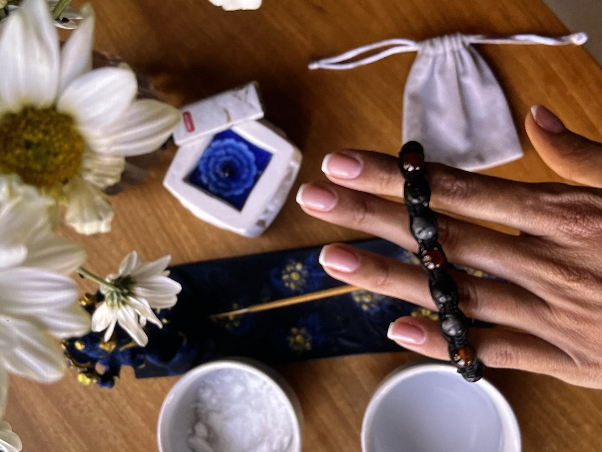 The image depicts a close-up, overhead shot on a wooden surface. A hand with manicured nails holds a black beaded bracelet with alternating red and black beads. To the left, white daisies are visible, along with a small white container with a blue floral design, and a dark blue and gold patterned object with an incense stick on top. Two small white cups sit at the bottom of the frame. In the upper right is a small, drawstring white pouch. The city of Unknown, Indonesia, cannot be determined from the image.