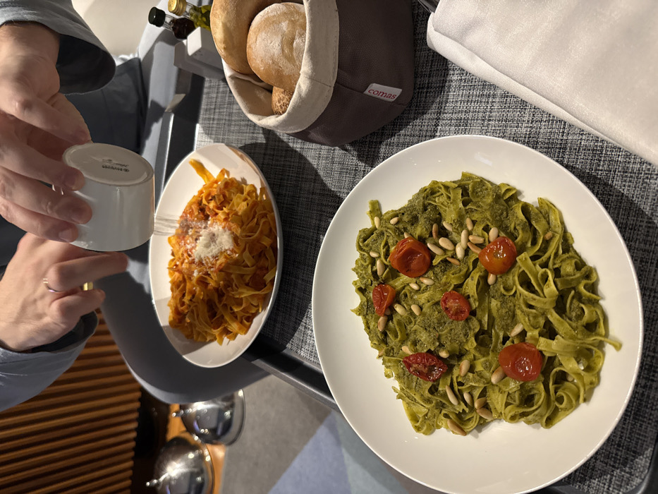 Two plates of pasta sit on a table. One plate holds pasta with a red sauce topped with grated cheese, a hand is adding it. The other has pasta with green pesto, topped with pine nuts and tomatoes. Bread rolls in a brown fabric basket are near the pasta. Bottles of olive oil are also visible.