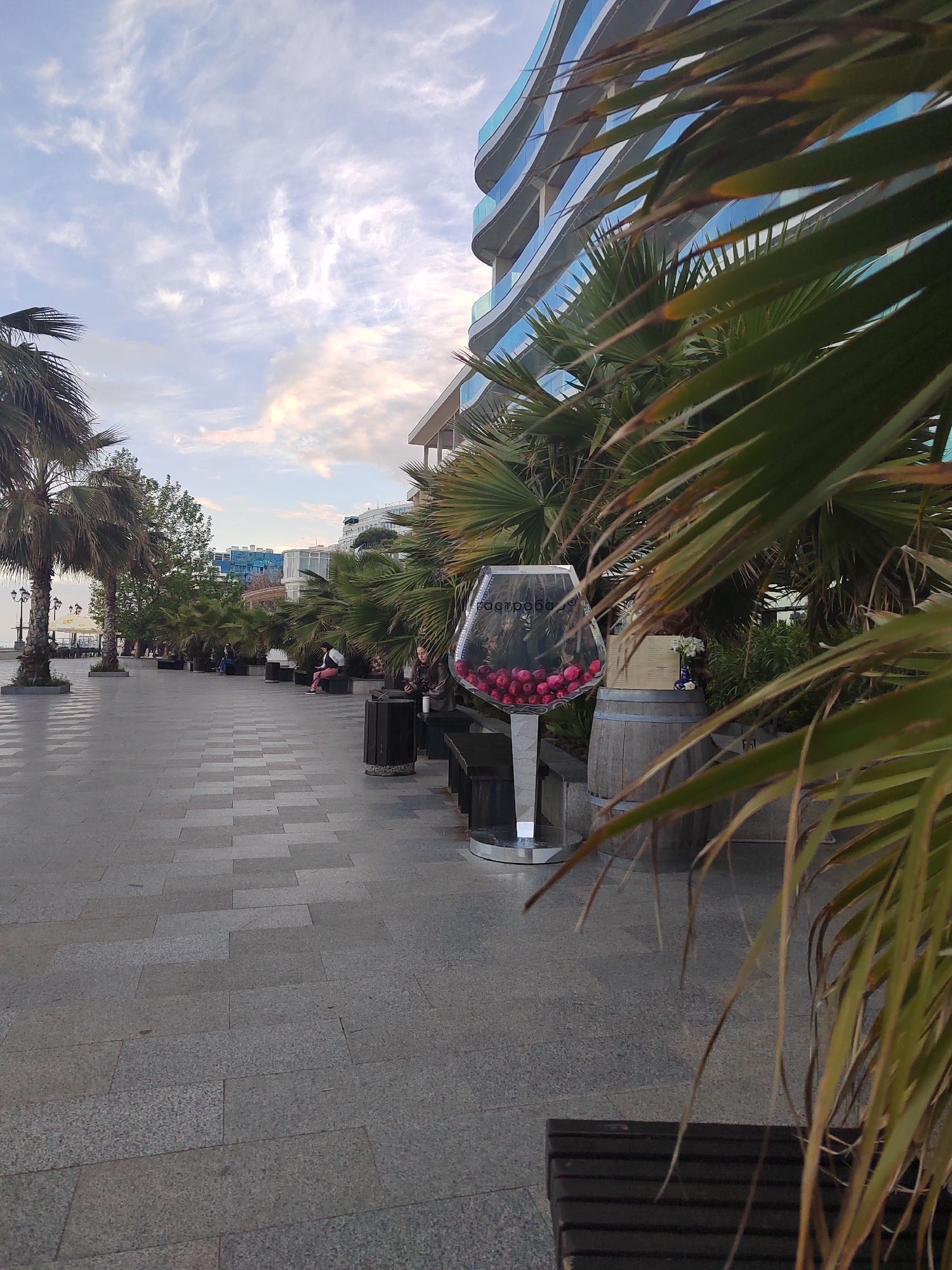 The image shows a promenade in an urban setting, with a gray-paved walkway lined with benches, palm trees, and a building with curved balconies. People are seated on benches along the walkway. A large, decorative wine glass filled with red objects stands near a wooden barrel. The sky is partly cloudy. The location appears to be in the city of Unknown, Unknown.