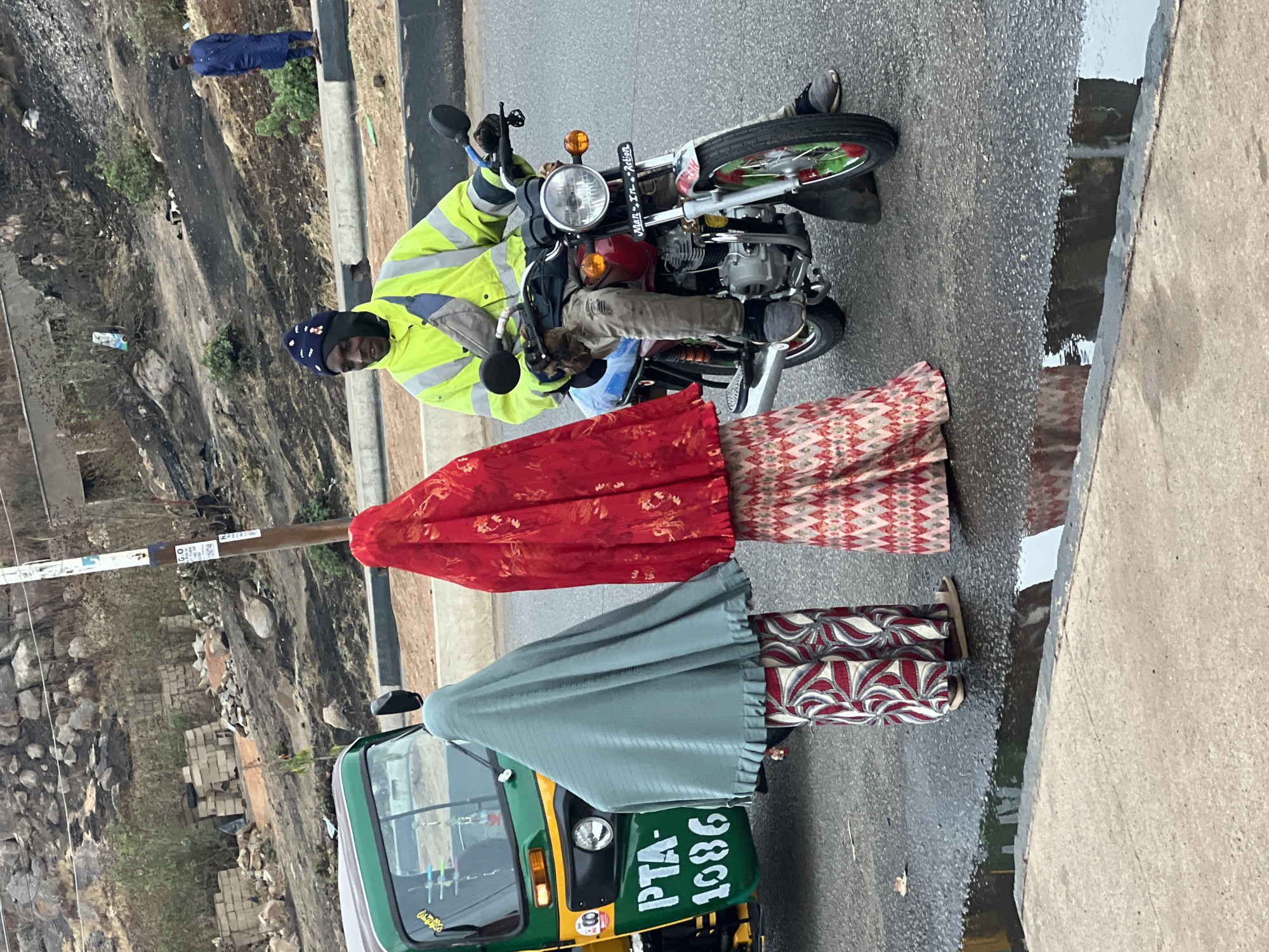 The image depicts a street scene in Jos, Nigeria. A man wearing a yellow reflective vest and a black cap is seated on a motorcycle parked on the road. Two women, largely obscured by a red and a green shawl, respectively, stand beside the motorcycle. The women are wearing patterned pants. In the background, there is a green auto-rickshaw with the license plate "PTA-1686". A concrete curb runs along the right side of the frame, with a pool of water on the road beside it. A steep embankment covered in vegetation and debris rises on the left. Another person wearing a blue outfit is on the slope in the distance.
