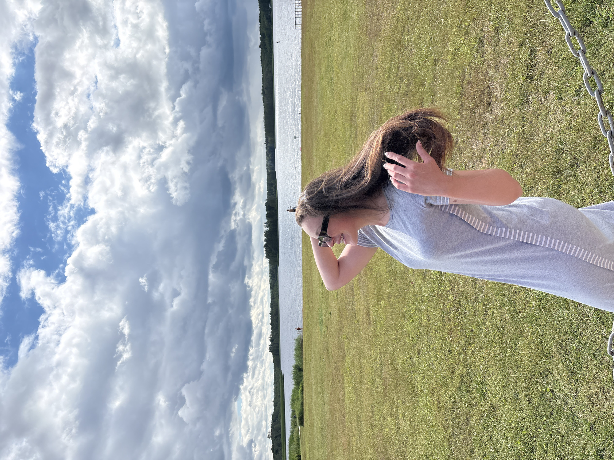 In Мазалаўскі сельскі Савет, Belarus, on a cloudy day, a young woman with long brown hair and sunglasses stands in a grassy field. She is wearing a grey dress with white stripes on the side. Her right hand is raised and touching her hair, which is blowing in the wind. In the background, there is a large body of water, likely a lake, with a treeline on the opposite shore. Several small figures are visible in the water and on the shore, suggesting a recreational area. A metal chain barrier is visible in the foreground, partially obscuring the bottom right corner of the image.