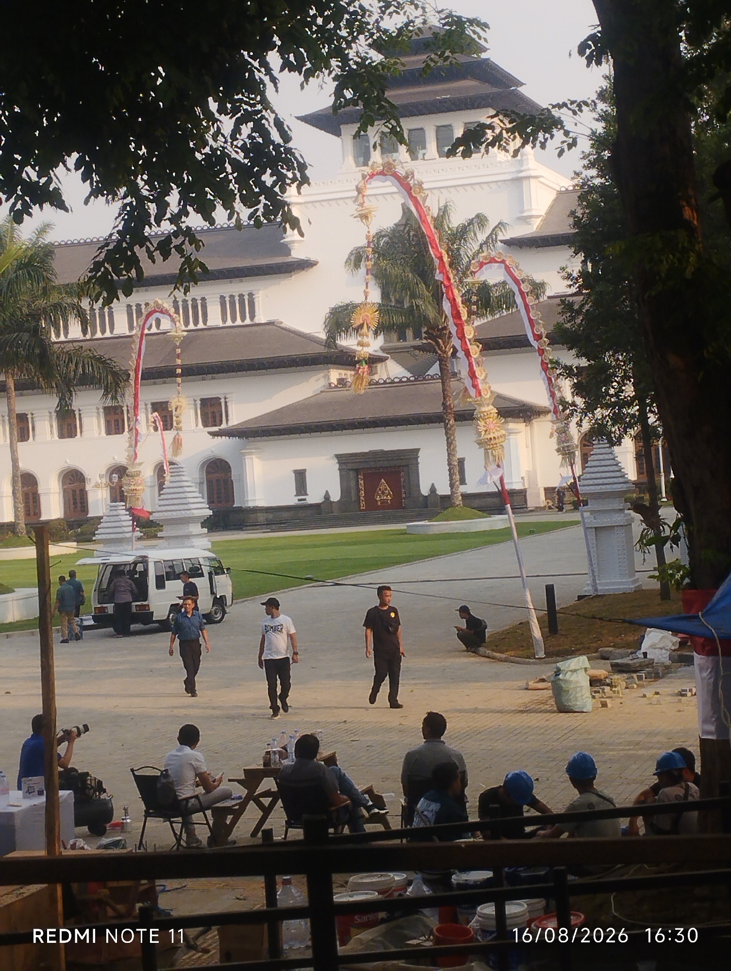 The image depicts a wide-angle view of the Gedung Sate in Bandung City, Indonesia. The building, a prominent landmark with distinctive architecture featuring tiered roofs and white facades, is visible in the background. In the foreground, several people are gathered in an outdoor area. Some individuals are standing near a white van, while others are seated at tables. A photographer is visible on the left, holding a camera. Various objects, including what appear to be construction materials and tarps, are scattered on the ground. The scene is set during daylight hours, with the sky appearing hazy. The overall impression is one of a public space, possibly a gathering or event, in front of the historical building.