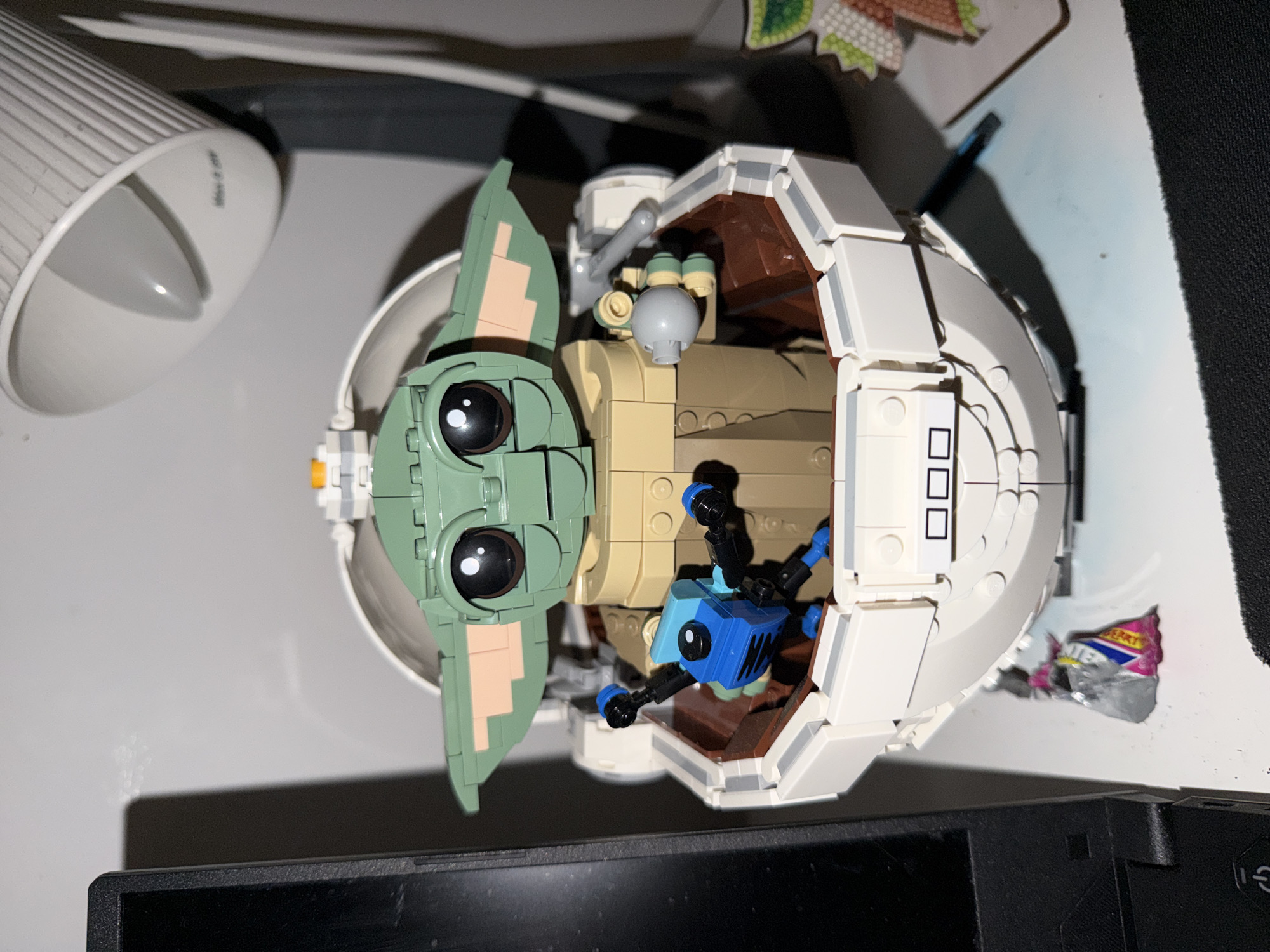 The image shows a Lego model of Grogu (Baby Yoda) in his hover pram, resting on a surface that appears to be a desk or table. A white lamp is in the upper left corner of the image, and a black electronic device (possibly a monitor or laptop) is partially visible in the lower part of the frame. There is a candy wrapper near the right side of the model. The background is generally out of focus and indistinct. The location in Budva, Montenegro is not discernible from the visual information provided in the image.