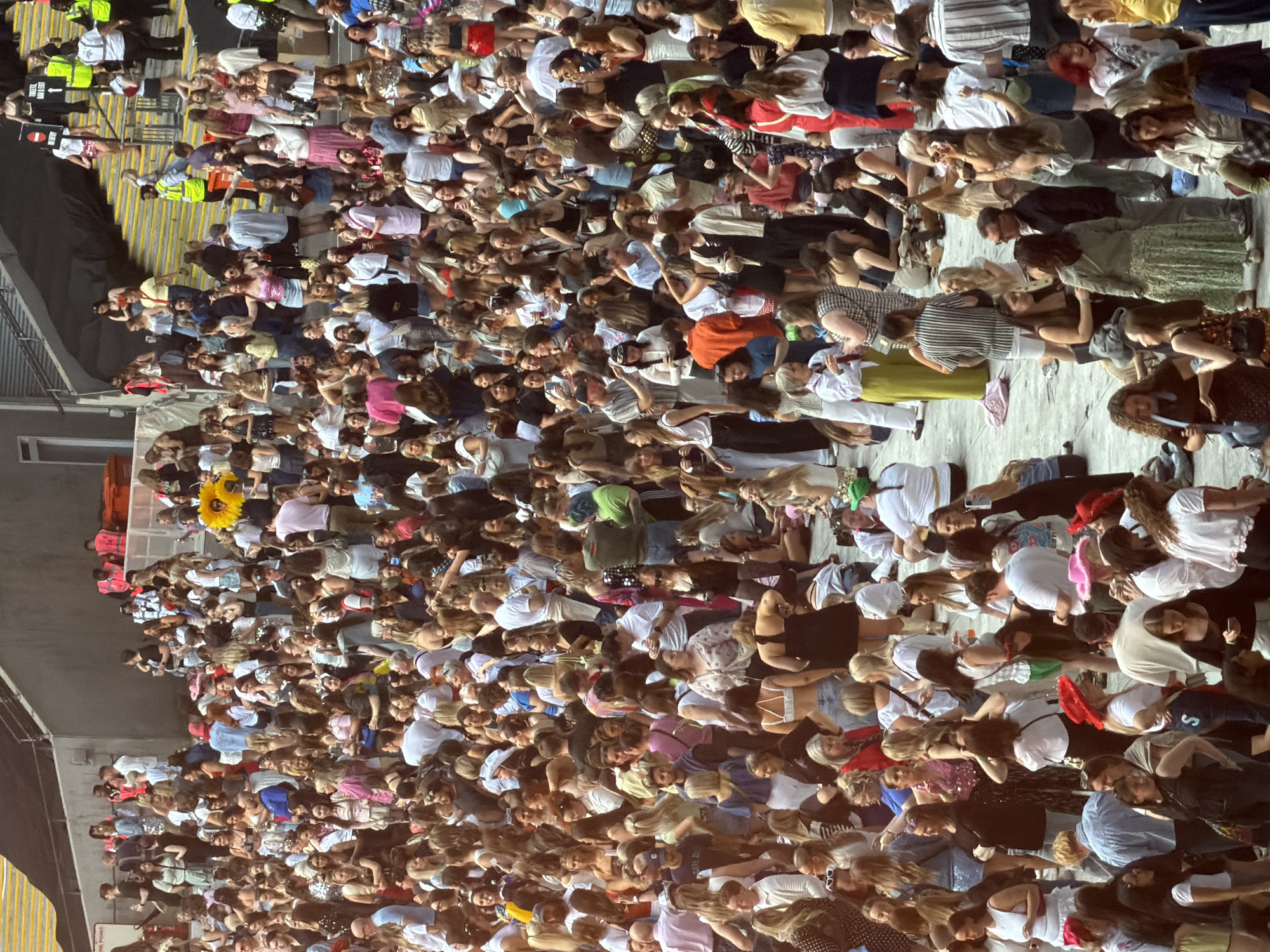 The image shows a large crowd of people gathered in an outdoor venue, likely a concert or festival, with stadium seating visible in the background. The crowd is densely packed, with individuals standing and sitting. Many people are dressed in casual attire, and some appear to be holding drinks or phones. The overall atmosphere seems lively, with a mix of ages and demographics present. The scene is set on a bright, possibly sunny day. No specific London landmarks are identifiable, but the architecture suggests a modern entertainment venue.