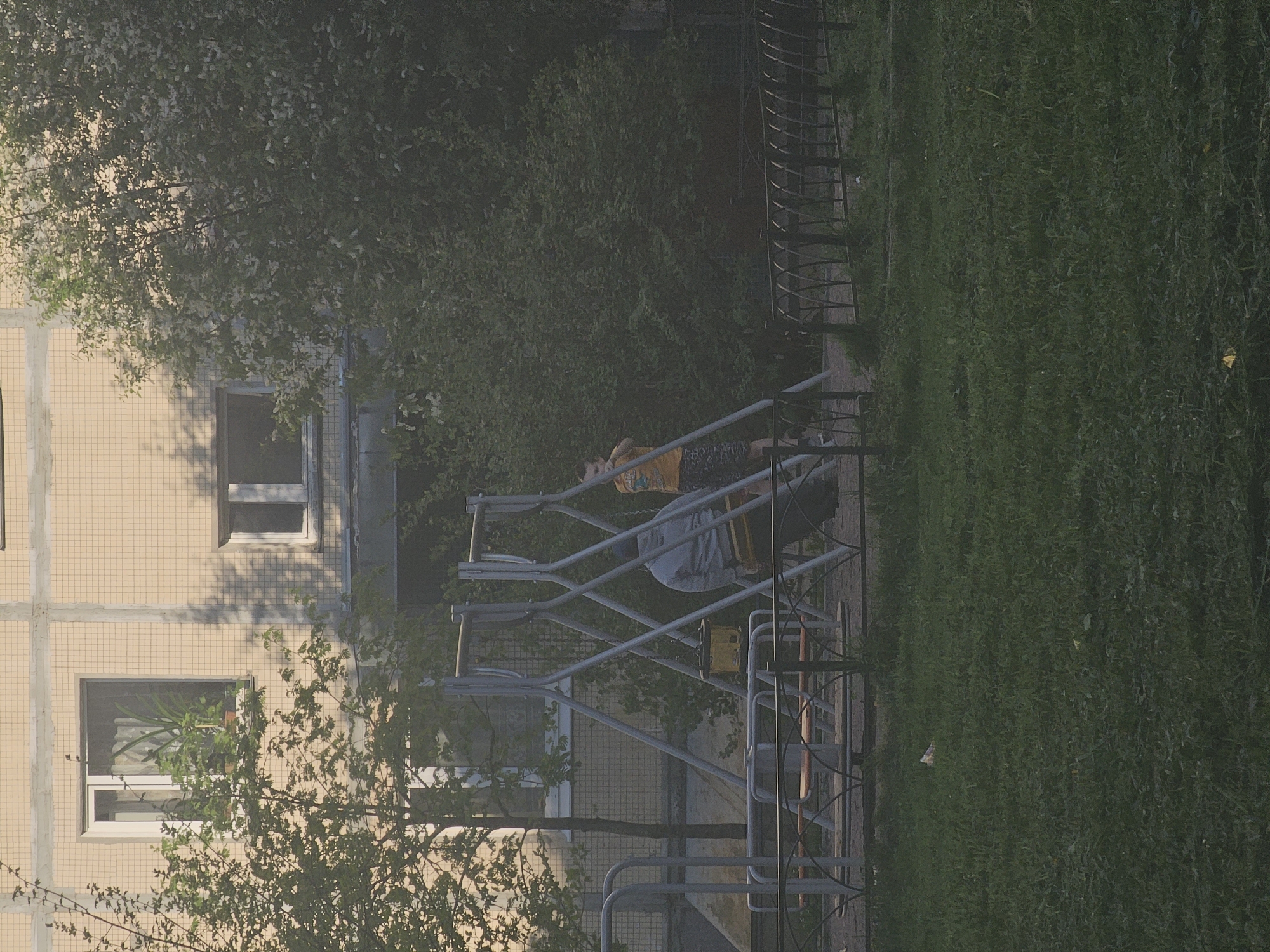 The image depicts a playground adjacent to a building in what appears to be Tosno, Russia. A person wearing a yellow shirt and patterned pants rests on playground equipment, specifically a seesaw. The playground sits on a grassy area, bordered by a fence. The building is tiled and features two visible windows; one has a potted plant inside. Trees are present, partially obscuring the view of the building and playground.