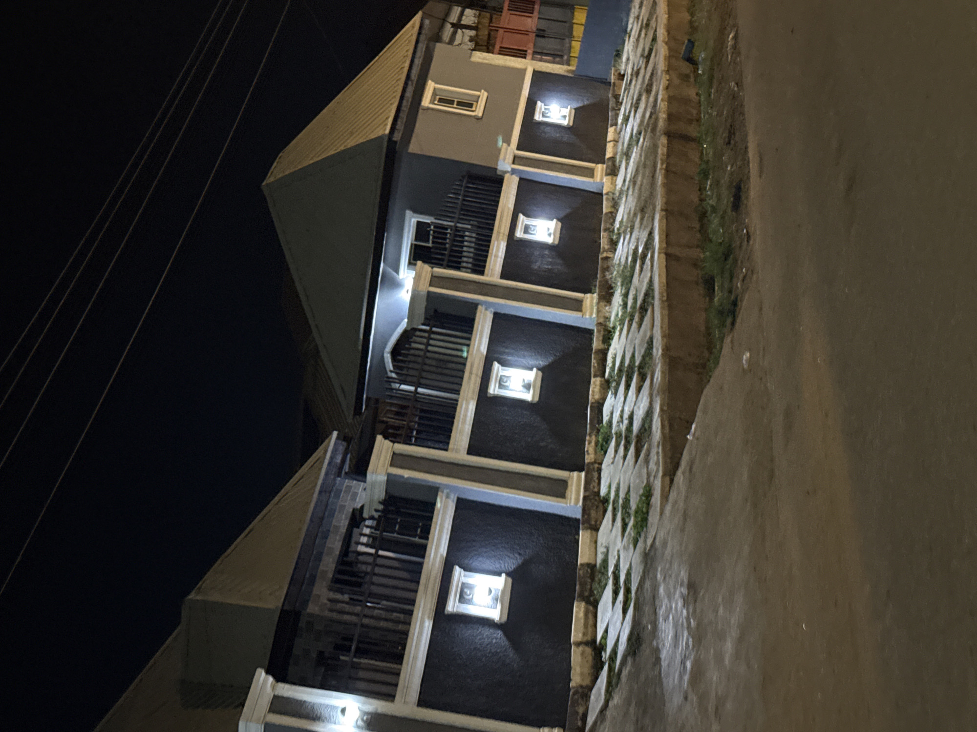 The image shows a nighttime exterior view of a two-story building, likely a residential property, in the city of Abuja, Nigeria. The building features a dark gray exterior with contrasting lighter gray and white trim. There are two prominent, pointed roofs, each with a metallic finish. The lower level of the building has several small, rectangular windows with internal lighting that casts a warm glow. The upper level has a balcony with black metal railings, accessible through windows. A paved walkway with square concrete slabs and patches of grass is visible in front of the building. The street in the foreground is unlit, and overhead power lines are visible against the dark night sky. The scene appears calm and devoid of human activity.