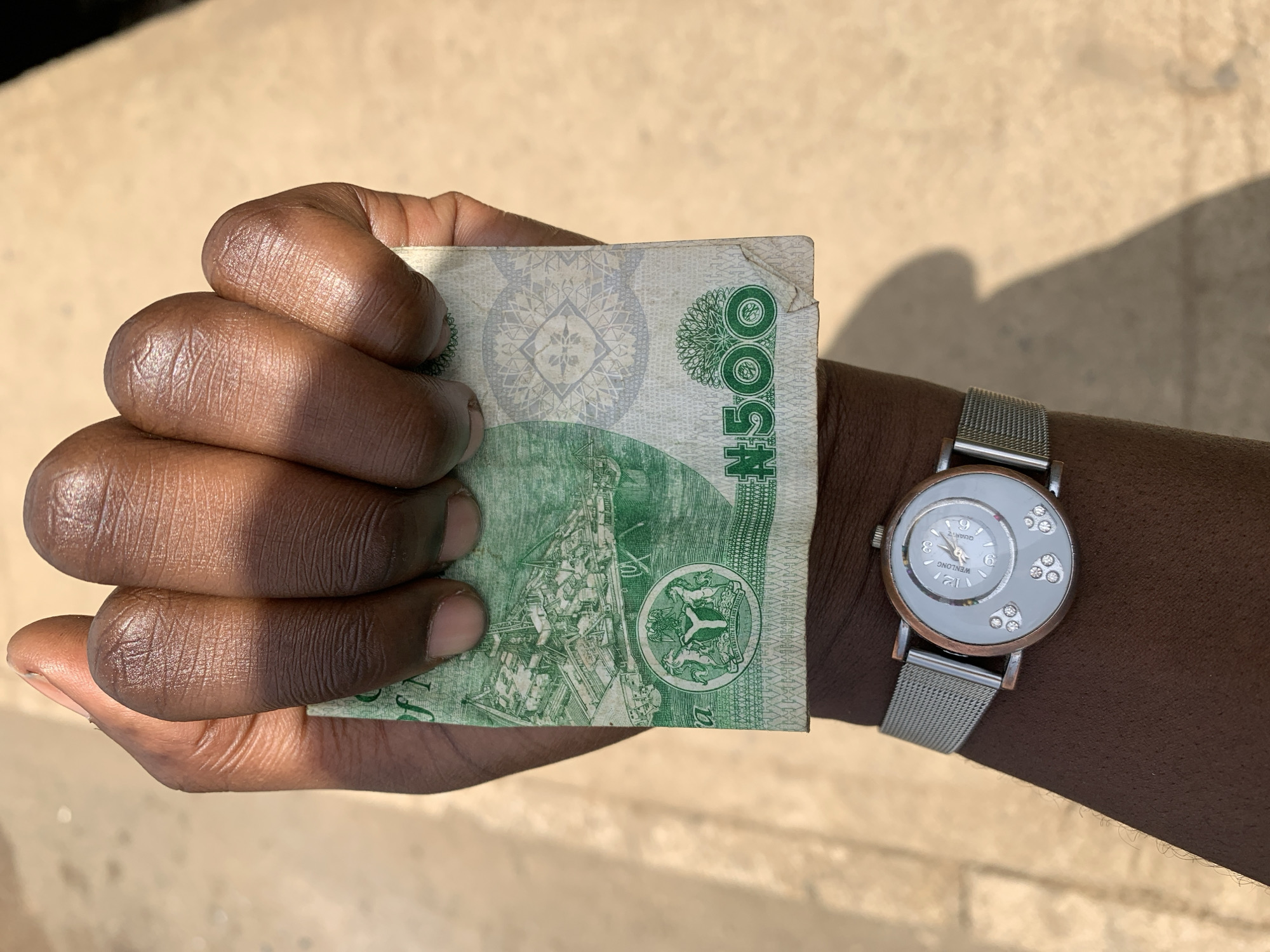 A close-up shot of a hand holding a Nigerian 500 Naira note in what appears to be Rafin Par, Nigeria. The hand is dark-skinned with visible wrinkles and holds the green banknote, which is partially crumpled. A wristwatch with a metallic strap is on the wrist. The background appears to be a wall or pavement. The environment is bright.