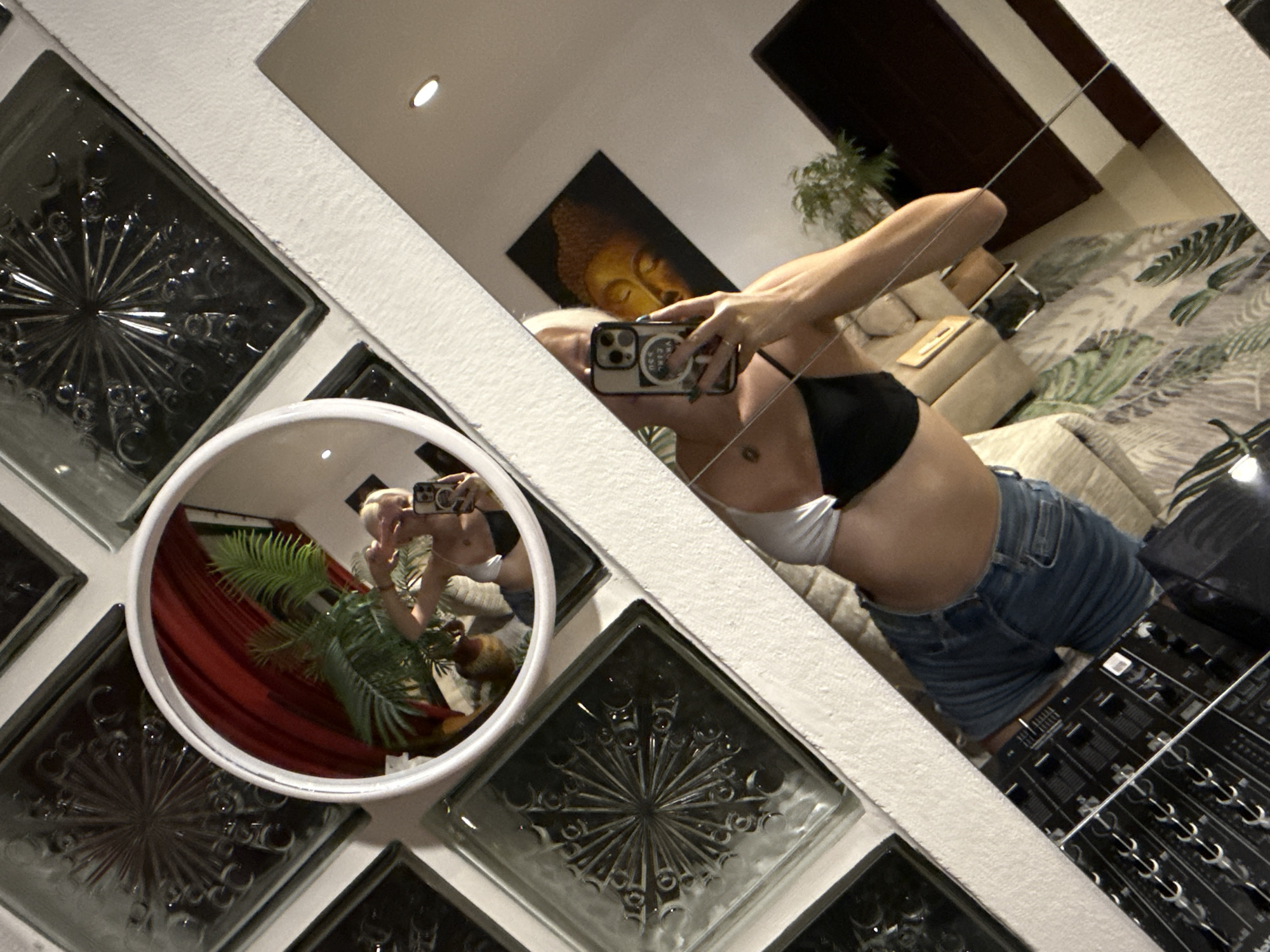 A woman with blonde hair is reflected in a mirror, taking a selfie. She is wearing a black and white bikini top and denim shorts. The mirror is angled, showing the woman's torso and arm as she holds her phone. Behind her, another mirror reflects a potted plant and a painting of the Buddha. In the foreground, a DJ mixer is visible. The room appears to be dimly lit, with decorative glass blocks on the wall. The overall impression is a casual, personal moment captured in a possibly tropical setting.