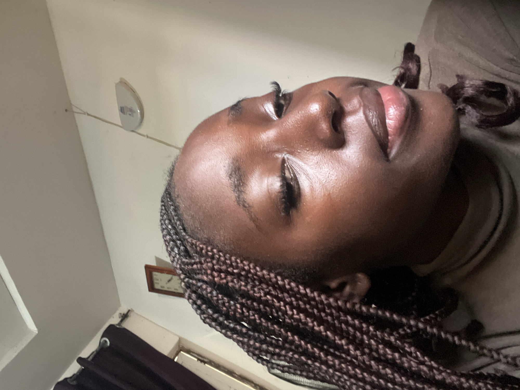 A medium shot depicts a Black woman with braided hair lying down. The camera angle is low, capturing her face from below. She has dark skin, full lips, and her eyes are closed. Her braids are brown and neatly arranged. In the background, a wall is visible with a light fixture and a clock. A dark curtain partially covers a window. The location of the photo is likely indoors, but specifics about Abuja, Nigeria, or a precise location are not discernible from the image alone.