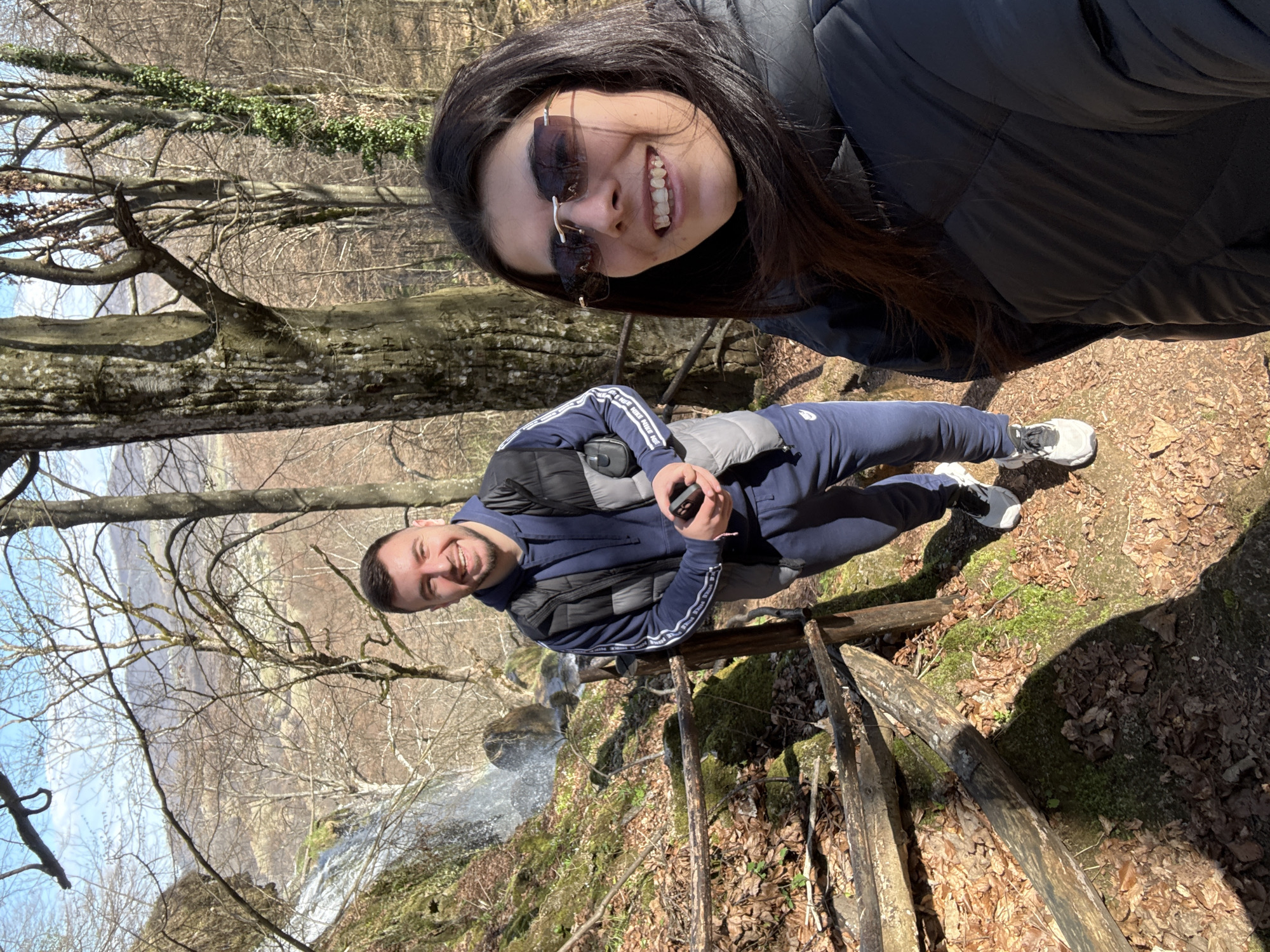 Two people are posing for a selfie near a waterfall. A woman with dark hair and sunglasses is smiling at the camera. Behind her and slightly lower is a man wearing a navy tracksuit and vest, also smiling. In the background, there is a waterfall with mossy rocks and bare trees. The scene appears to be a hiking trail with a wooden fence, likely in a wooded area. The image was taken in Ribaritsa, Bulgaria.