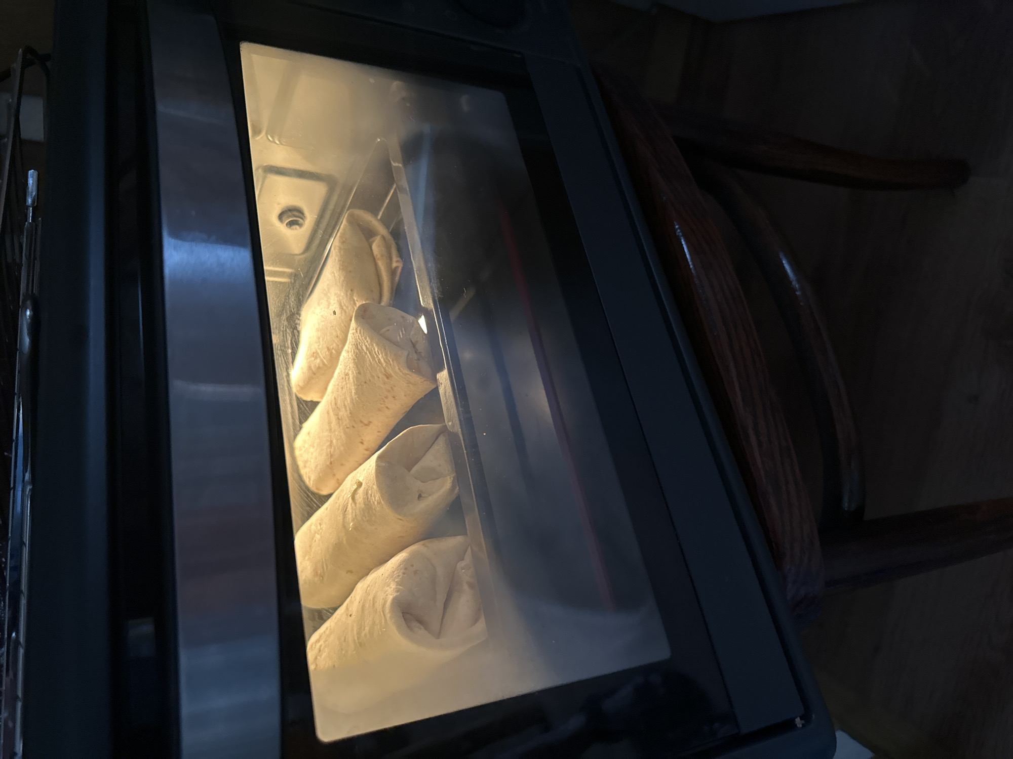 Four burritos are arranged in a toaster oven on a rack. The burritos appear to be uncooked and are tightly wrapped in a light-colored tortilla. The toaster oven is closed, and the interior light is on, illuminating the burritos. The background outside the oven is dark, showing hints of a wooden surface and chair legs, suggesting an indoor setting. No people are visible in the image. The location is described as Norviliškės, Lithuania.