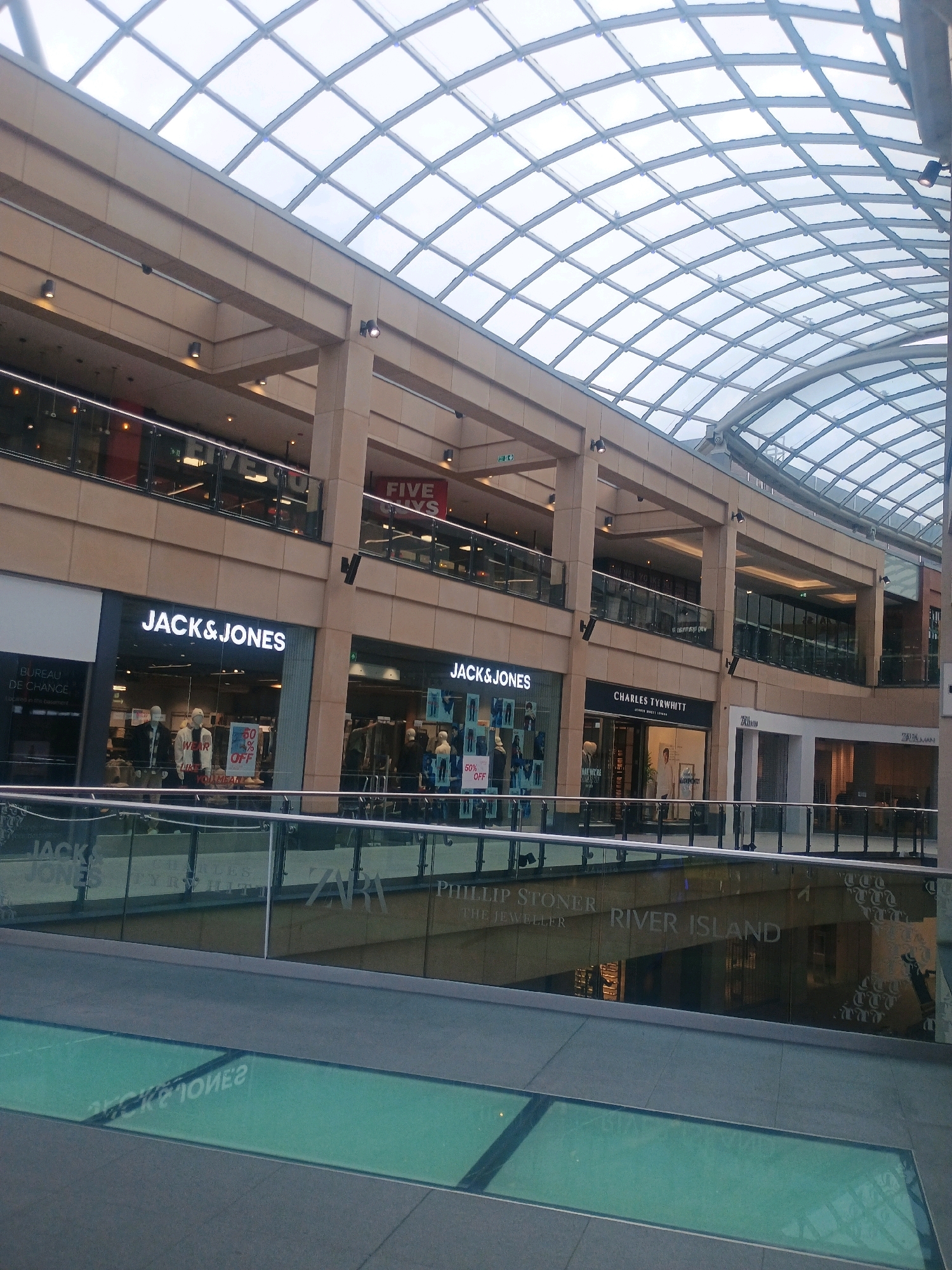 The image shows the interior of a modern shopping mall with a large, multi-level structure and a distinctive glass-panelled, arched ceiling. On the ground level, several retail stores are visible, including "JACK & JONES," "CHARLES TYRWHITT," "ZARA," and "RIVER ISLAND." The stores feature large display windows with mannequins and merchandise. A glass-panelled walkway or bridge with a railing is present on a higher level, allowing a view into the shops and across the mall. The overall environment appears clean and well-lit, suggesting a commercial and public space.