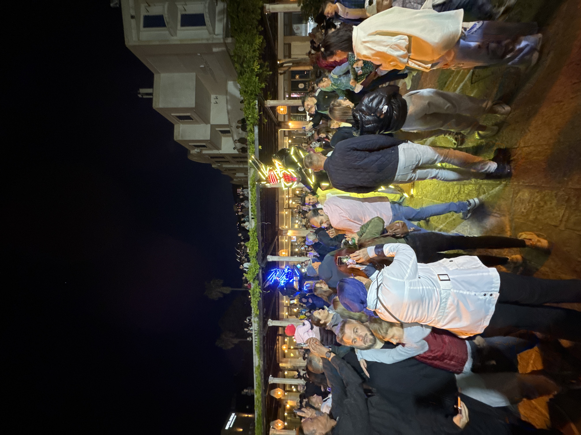 The image depicts an outdoor night scene featuring a large gathering of people, likely in Budva, Montenegro. The crowd, predominantly adults, fills the foreground and midground, oriented towards a central point on the left side of the frame. Many individuals are holding up smartphones, indicating they are either recording video, taking photos, or engaging with a performance or spectacle.
The atmosphere appears lively and celebratory, enhanced by festive lighting. In the midground, illuminated decorations are visible, including structures adorned with bright blue and red lights, one resembling a stylized heart. Overhead lighting fixtures or lanterns provide additional illumination, casting a warm glow over the scene.
On the left side of the image, under a dark night sky, several light-colored buildings with windows are visible, suggesting an urban or resort environment. Green foliage, possibly trees or hedges, lines the area where the people are gathered and extends into the background. In the far distance, a sign partially reads "HOTEL S," which, in the context of Budva, likely refers to Hotel Splendid. The scene captures a vibrant public event unfolding on a clear night.