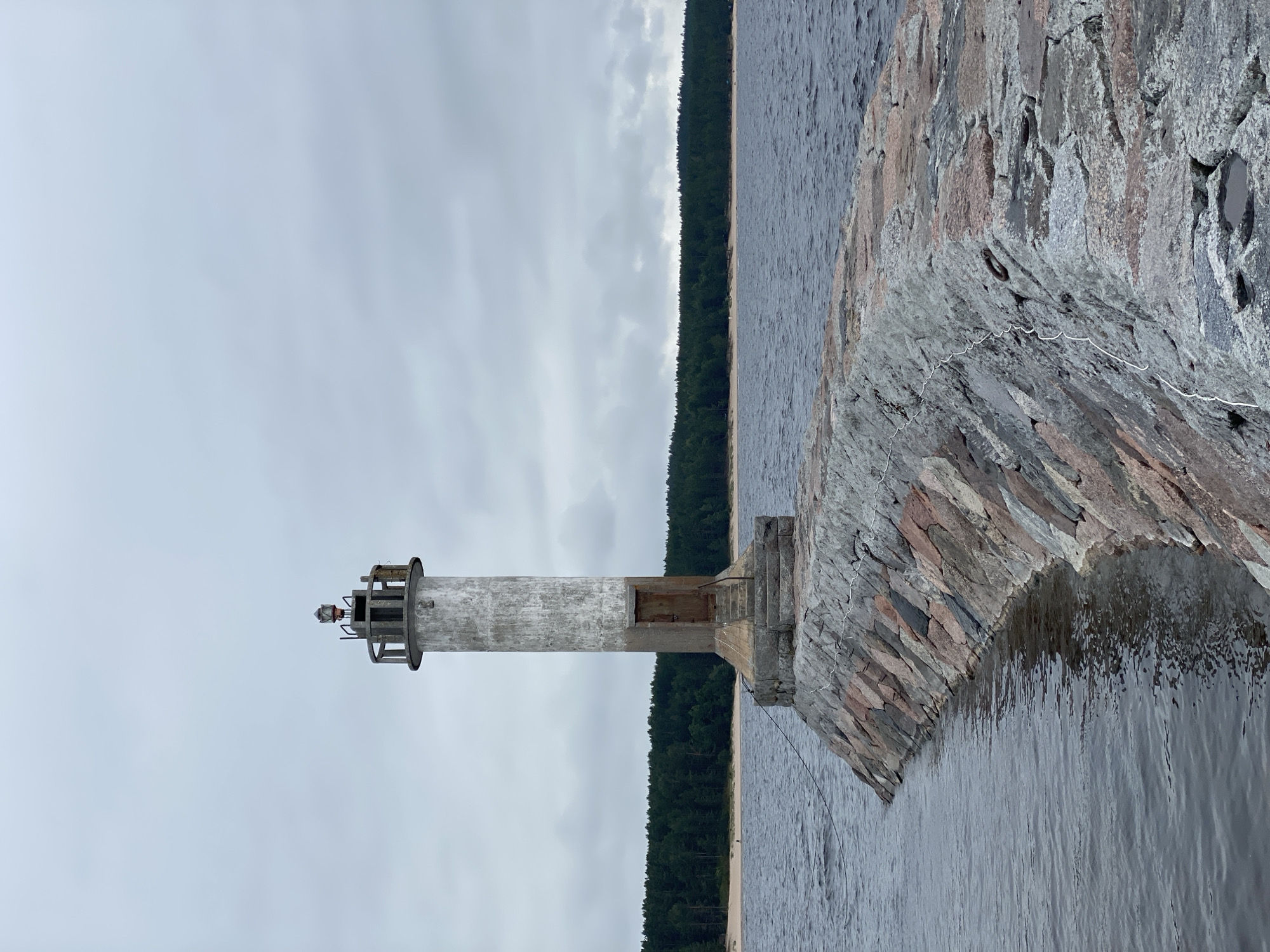 The image shows a concrete lighthouse situated on a stone pier extending into a body of water under an overcast sky. The lighthouse is cylindrical with a weathered, grey exterior and a metal railing structure at its top, which houses the lamp. A rusty metal door is visible on the lower part of the lighthouse, accessible by a short flight of stone steps. The pier is constructed from large, irregularly shaped stones of varying colors, including grey, pink, and reddish hues, and is lined with what appears to be white rope or cable. The water is dark and choppy, with ripples indicating a slight breeze. In the background, a dense forest of green trees lines a sandy beach. The overall scene suggests a remote coastal or lakeside location. The provided crop hints that the lighthouse may be located near Моторное, Russia.
