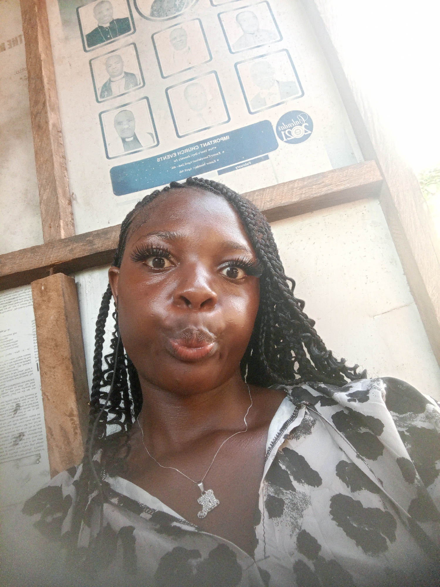 A woman with braided hair stands in front of a notice board in Mbribit, Nigeria. The notice board features portraits of several men, possibly religious figures. The woman wears a light-colored, patterned blouse and a silver necklace. She is looking at the camera with pursed lips. The background suggests an outdoor or semi-outdoor setting.
