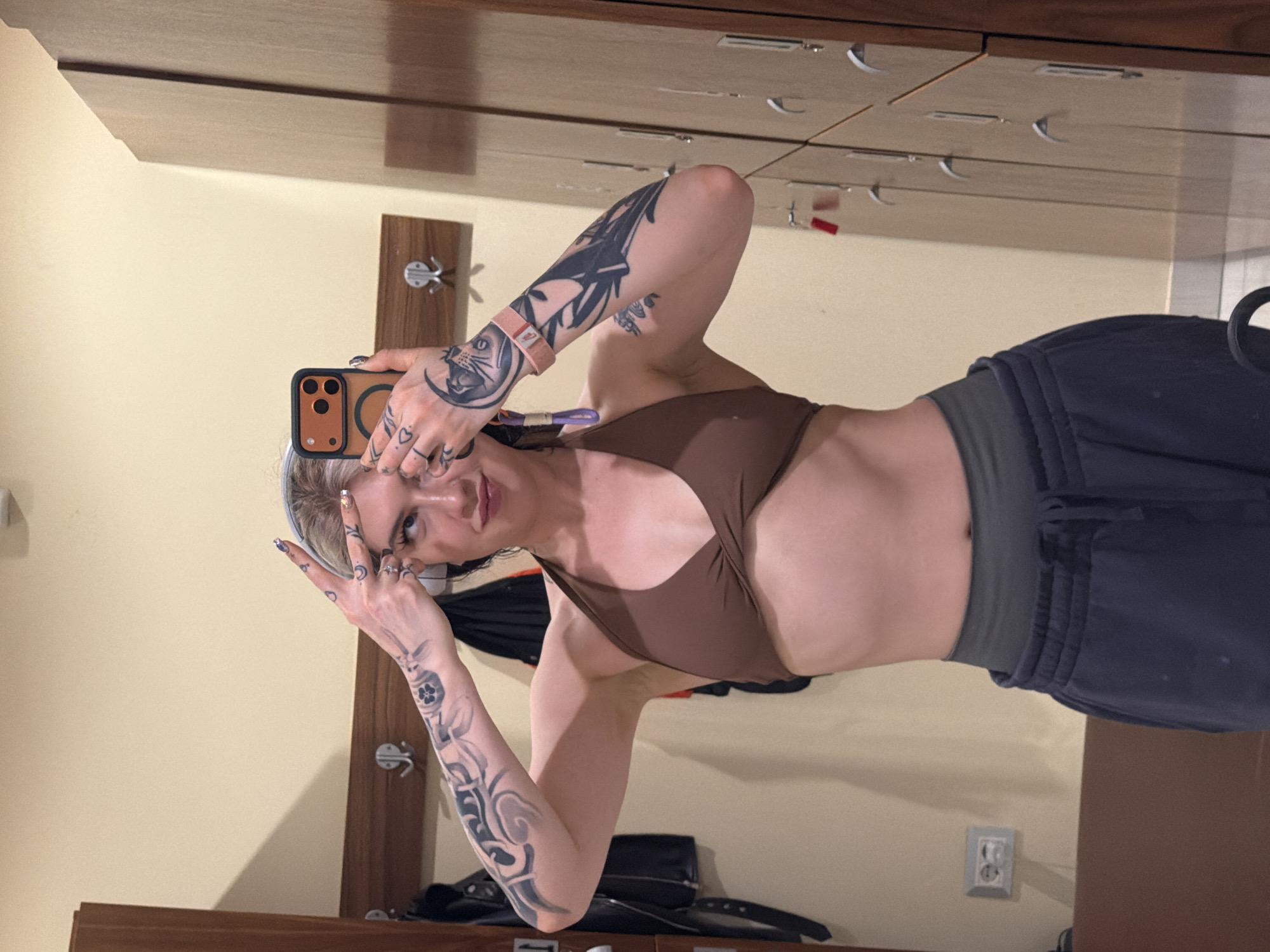 A person with tattoos on their arms is taking a selfie in front of a mirror in a locker room. They are wearing a brown top and dark pants. The locker room has light yellow walls, brown lockers, and metal hooks. An electrical outlet is visible on the wall near the bottom right corner. A black jacket and other items are visible on the locker shelves. There is no specific information that would allow for the determination of a precise location within the city of Budva, Montenegro.