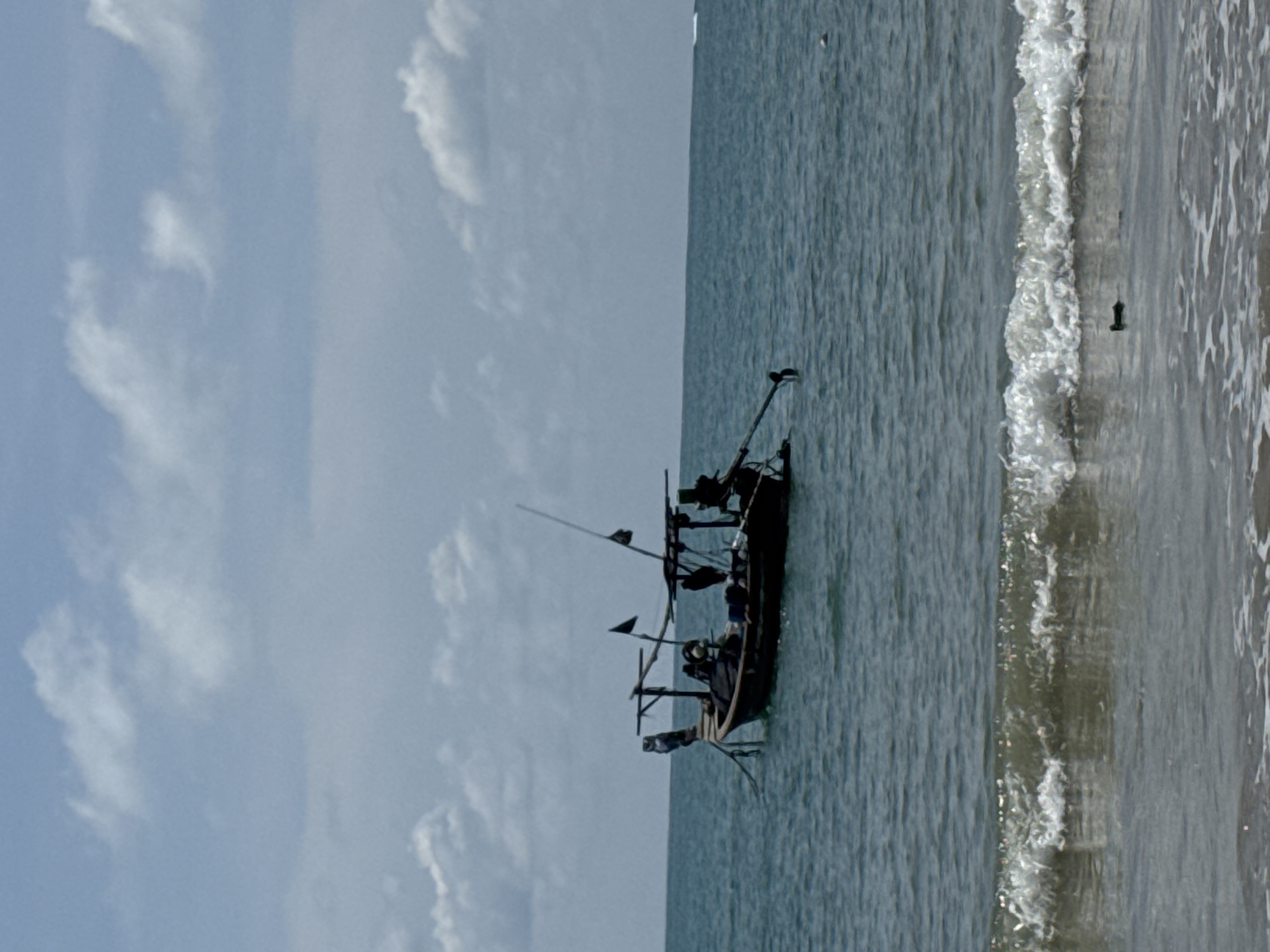 A long, narrow wooden fishing boat with a dark hull is anchored in the sea off the coast of Sakhu, Thailand. The boat has a canopy structure and two masts, one with a black flag. Two figures are visible on the boat. The water is choppy, with small waves breaking in the foreground. The sky is cloudy, with patches of blue. The scene appears to be during daylight hours.