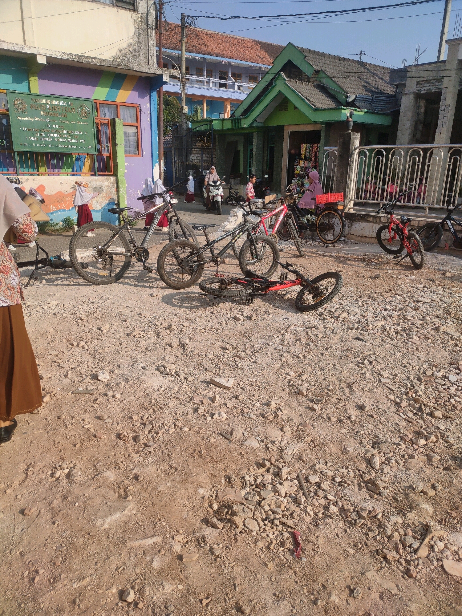 In Payaman, Indonesia, on a sunny day, a street scene unfolds with a colorful building in the background, possibly a school, adorned with bright murals and a green sign with Indonesian text. A few children in school uniforms, including hijabs, are visible near the building and by parked motorcycles. The foreground is dominated by a dirt and gravel ground with several bicycles, some upright and some lying on their sides. One red bicycle in the foreground is on the ground, its wheels pointing in different directions. Further back, other bicycles, including a red one and a black mountain bike, are parked or standing. A woman wearing a headscarf and a long skirt is partially visible on the left side of the frame, walking away from the viewer. The environment appears to be an outdoor area in front of buildings with a fence in the middle ground.