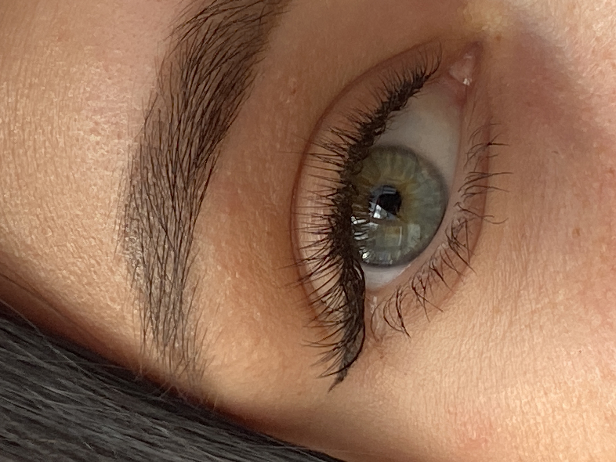 A close-up, high-resolution image depicts the left eye and eyebrow of a person, observed from a slightly elevated and side angle. The eye features a striking green-grey iris with a visible dark pupil, reflecting a subtle light source. The eyelashes are long, dense, and dramatically curled upwards, indicative of professionally applied eyelash extensions, which are dark black and voluminous.
Above the eye, the eyebrow is full, dark, and appears well-groomed, with individual hair strands clearly visible. The skin surrounding the eye and eyebrow exhibits natural texture, including visible pores and slight variations in tone, particularly around the brow bone and the inner corner of the eye, where some minor redness or texture is present. The lighting is soft and even, highlighting the intricate details of the eye and its enhanced features. The image captures a static moment, focusing entirely on the eye. No specific activity or event is taking place, and no text is visible within the frame. The location context for this media is Chaikovsky, Russia.
