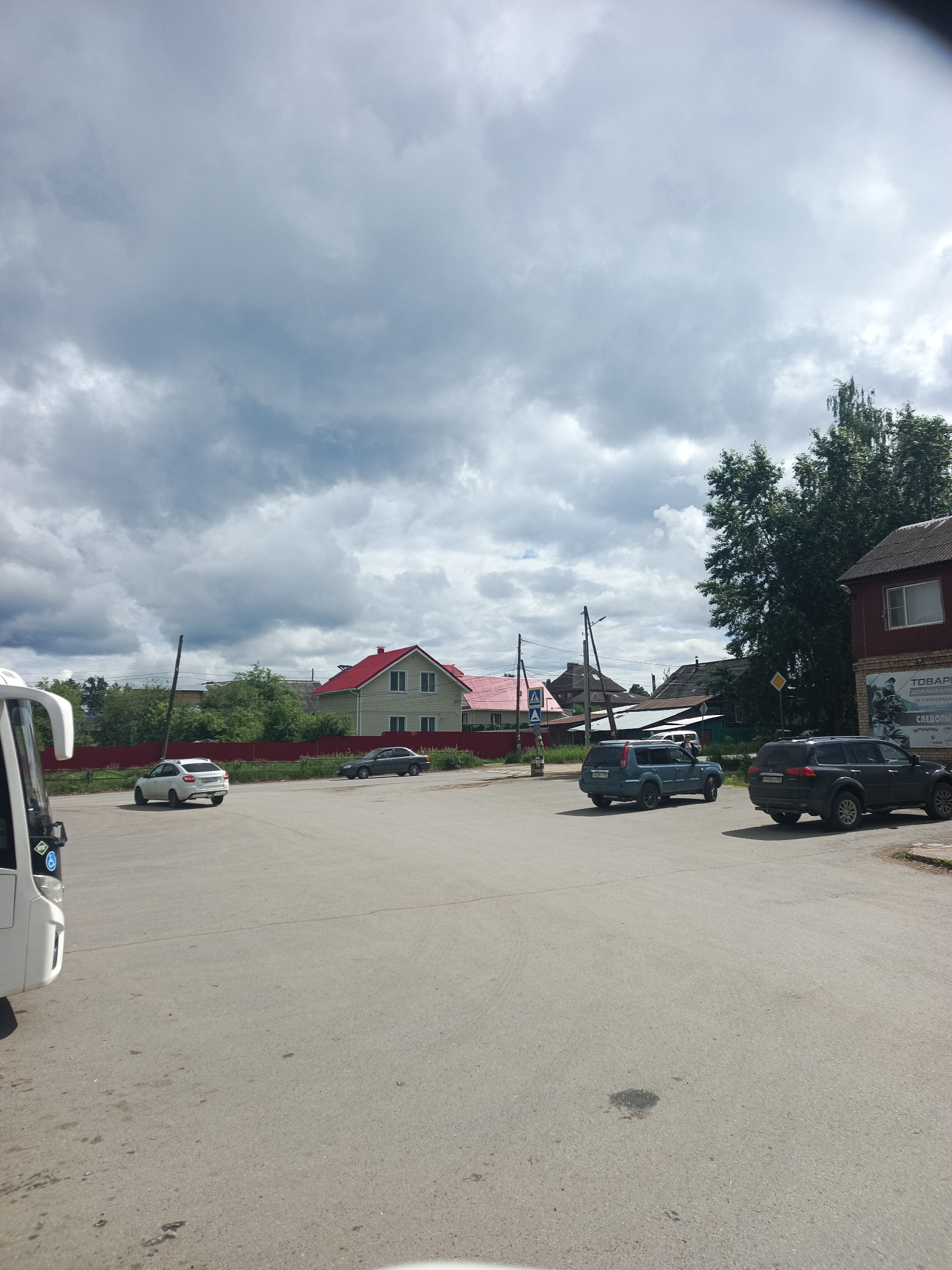 The image captures an outdoor scene, likely a parking area or wide street in a semi-urban or rural setting, under a heavily overcast sky. Thick, grey clouds dominate the upper portion of the frame, suggesting a cloudy day or impending rain.
In the immediate foreground on the left, the front-left corner of a white bus or large van is visible, featuring a blue accessibility sticker on its side. The ground consists of a large, grey paved surface, possibly asphalt, with some minor stains and irregularities.
Several vehicles are present in the midground and background. A white hatchback, possibly a Lada Granta, is seen from the rear, positioned left of center. Further back, a dark grey sedan is partially visible. To the right, a dark blue SUV, resembling a Nissan X-Trail, is parked or moving towards the left. Behind it, a dark grey Mitsubishi Pajero Sport SUV is seen from the rear-right, also parked or slowly moving.
The area is flanked by residential buildings and abundant greenery. On the left, behind a red fence, stands a light green house with a prominent red pitched roof and white-framed windows. Further in the background, more houses with varying architectural styles and roof colors (including more red roofs) are visible amongst numerous green trees. Utility poles with overhead wires are scattered throughout the scene.
On the right side of the image, a reddish-brown building features a dark roof and a large sign on its side. Visible Cyrillic text on the sign reads "ТОВАРЫ ДЛЯ РЫБАЛКИ И ТУРИЗМА" (Goods for Fishing and Tourism), with "СЛЕДОПЫТ" (Pathfinder) below it, and "YANAN" also visible. A yellow diamond-shaped road sign (indicating a priority road) is mounted on a pole near this building, and a blue pedestrian crossing sign is visible further left.
The time of day is daytime, with soft, diffused light due to the heavy cloud cover. The Cyrillic text and general appearance of the buildings suggest the location is in Russia, consistent with the provided context of Chaikovsky. There is no explicit human activity visible, giving the scene a quiet and static feel.