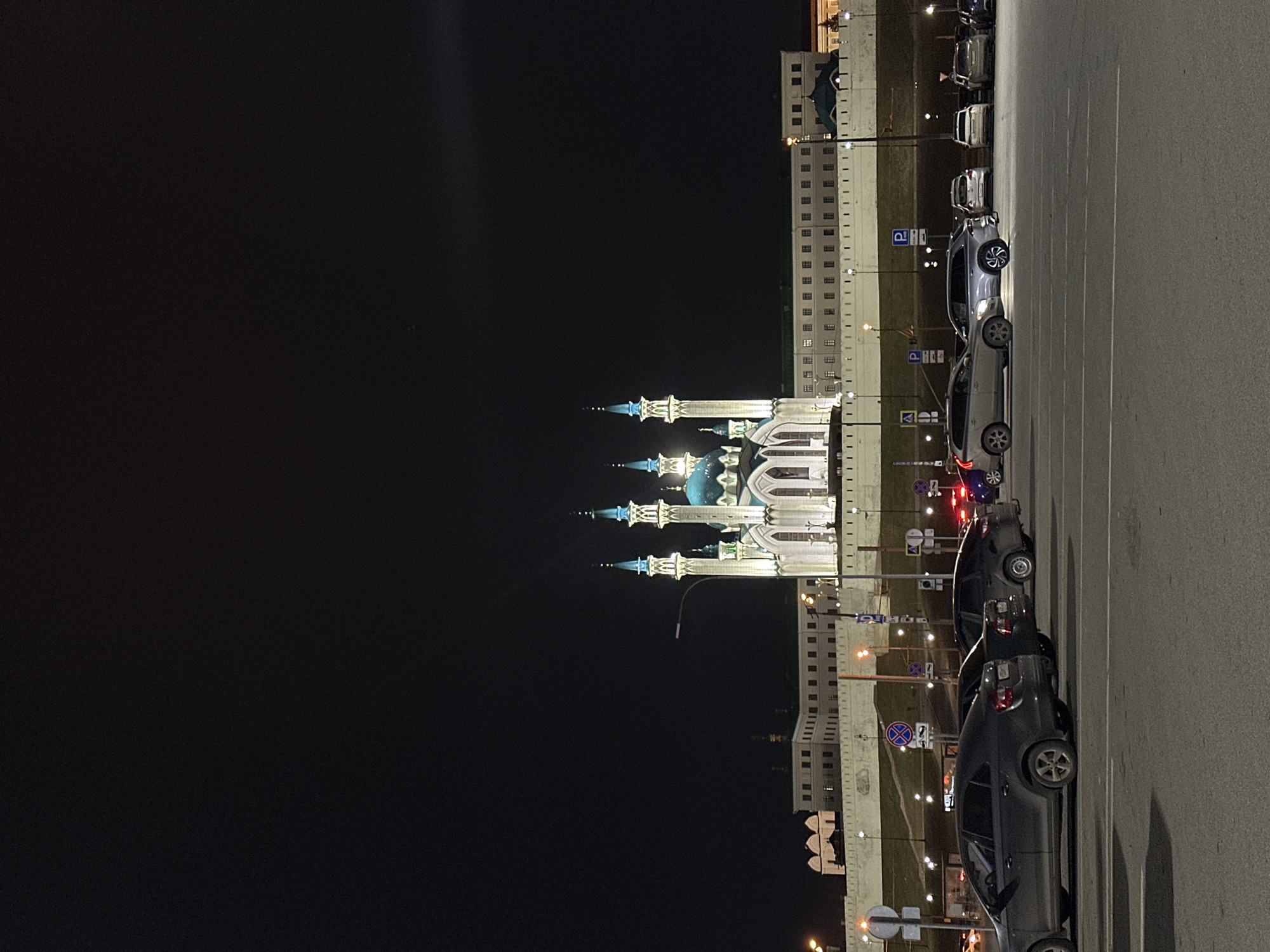 The image depicts the Qolşärif Mosque in Kazan, Russia, at night. The mosque's illuminated blue domes and minarets are visible against the dark sky. Adjacent to the mosque, a light-colored building with multiple windows is present. In the foreground, a parking lot is visible with several cars parked in designated spaces. Street lights illuminate the parking area and the surrounding buildings. Signage and traffic-related elements can be seen along the perimeter of the parking lot.