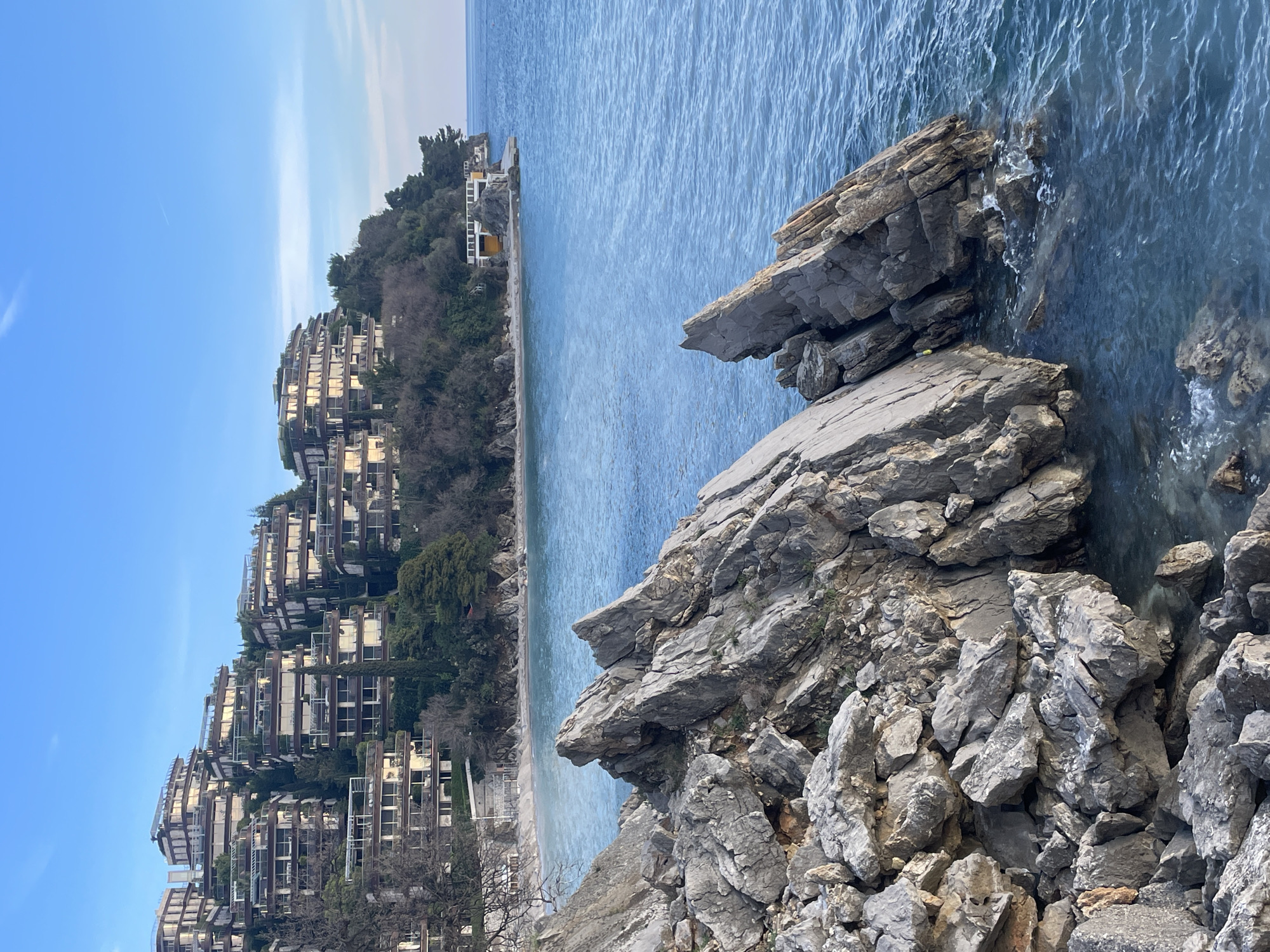 The scene depicts a coastal landscape in Budva, Montenegro, specifically the area around Dukley Gardens. In the foreground, a prominent cluster of jagged, grey rock formations rises from the lower left, extending into the calm, clear blue-green waters of the Adriatic Sea. The rocks exhibit fractured surfaces and appear partially submerged, with minor splashes and ripples visible where they meet the water.
In the midground, a crescent-shaped sandy or pebbly beach is visible, bordered by the sea and a steep, verdant hillside. To the right of the beach, a small, white-railed structure with a yellow base is positioned on a rocky promontory jutting into the sea. The water surface shows subtle variations in color, transitioning to a deeper blue further from the shore. No individuals are present.
The background is dominated by a densely vegetated hillside rising sharply from the coastline. Numerous modern, multi-story residential or hotel buildings are terraced into this slope. These structures feature flat roofs, extensive balconies, and large windows that reflect the ambient light, indicating a contemporary architectural design. The sky above is clear and light blue, with a faint, thin contrail visible in the upper expanse. The overall lighting suggests daylight conditions, possibly morning or late afternoon.