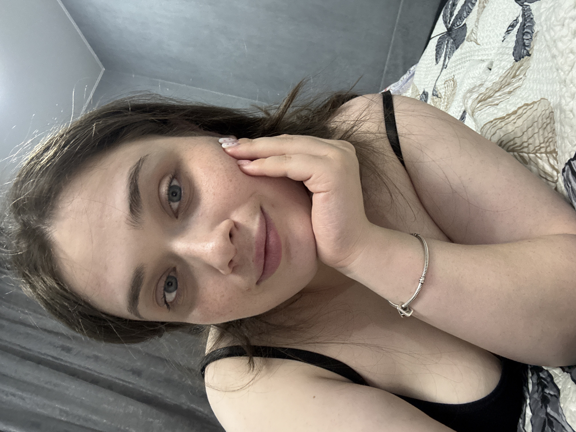 A woman lies on a bed, propping her face with her hand. She has light skin, brown hair, and blue eyes, with some freckles on her face. She wears a black tank top and a silver charm bracelet. The bed has a patterned bedspread. The background includes a gray wall and corner of the room. The location is Chaikovsky, Russia.