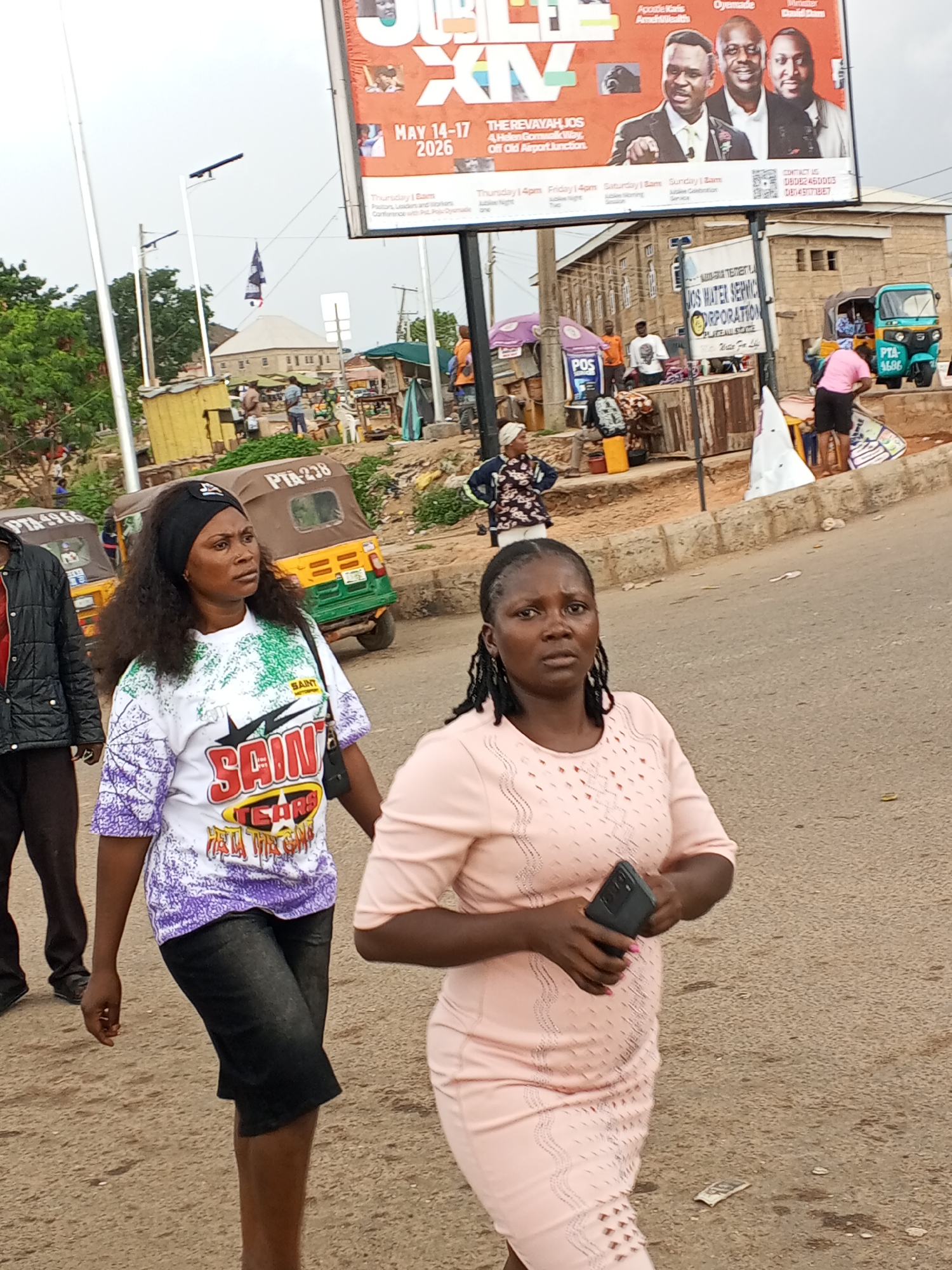 Here's a detailed description of the image:
**Setting:** An outdoor street scene in Gwong, Nigeria. The setting appears urban or peri-urban, featuring roads, pedestrian areas, and a variety of structures and vehicles.
**People:**
* Two women are in the foreground. The woman on the left wears a white t-shirt with the word "SAINT" on it, black shorts, and a black headband. The woman on the right wears a light pink dress and holds a smartphone.
* Several other people are visible in the background, including individuals near roadside stalls, a woman standing with her hands on her hips, and people near "POS" stands.
**Objects:**
* A large billboard advertises "THE REVAYAH, JOS," an event scheduled for May 14-17, 2026. It includes photos of men and contact information.
* A sign indicating "JOS WATER SERVICE CORPORATION."
* Several "PTA-238" and "PTA-4686" motorized tricycles (tuk-tuks) are present.
* Roadside stalls or vendors.
* A pole with a flag or banner.
**Actions/Interactions:**
* The two women in the foreground appear to be walking and engaged in a conversation.
* Other individuals are conducting business at stalls.
**Environment:** The weather is overcast or cloudy. A mix of informal vendor setups and infrastructure is present.