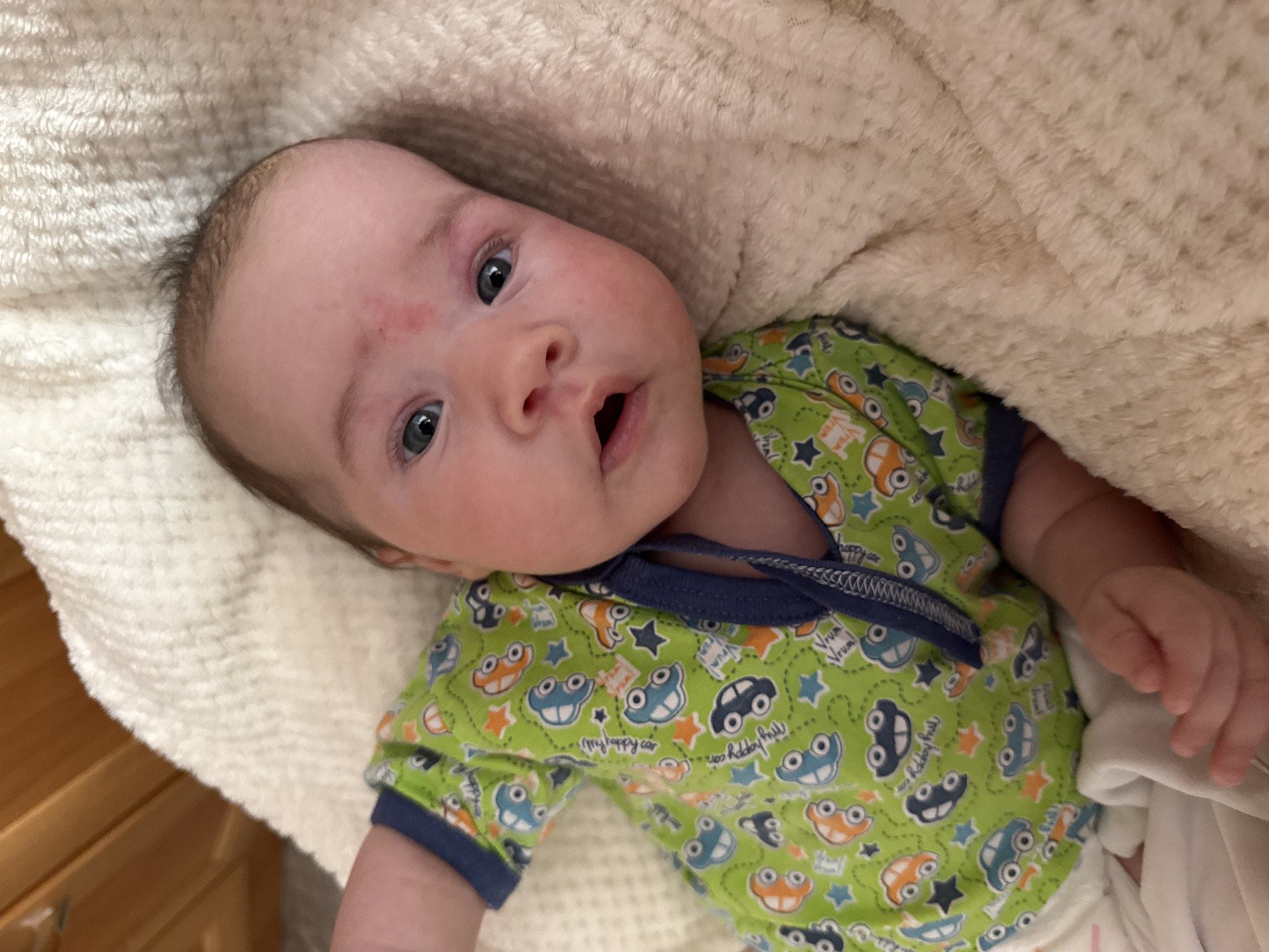 A newborn baby is lying on a cream-colored fluffy blanket, wearing a green onesie with a car print. The baby has a red mark on its forehead. The scene appears to be indoors, possibly in a nursery, given the presence of a wooden crib in the background. The lighting is warm and natural.