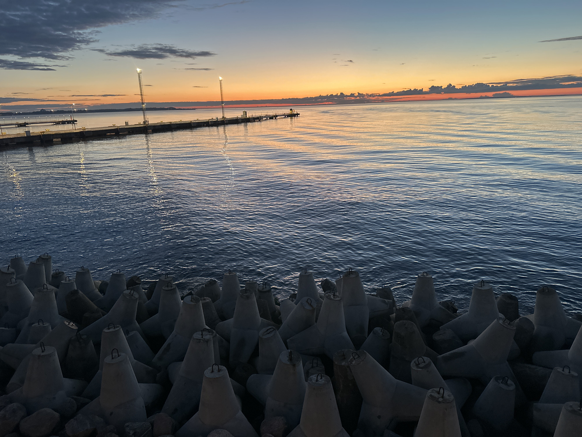 The image depicts a sunset over the Baltic Sea, viewed from a breakwater in Tallinn, Estonia. In the foreground, a dense arrangement of concrete tetrapods, commonly used for coastal protection, lines the edge of the water. These structures are grey and vary in size, with metal loops protruding from their tops. The sea is calm, reflecting the warm hues of the sunset. In the middle ground, a long pier extends into the sea, featuring several tall light poles with illuminated lamps. The sky transitions from a deep blue at the top to vibrant orange and pink near the horizon, where a distant landmass is visible. Clouds are scattered across the sky, catching the sunset's light. The overall scene is serene and showcases the coastal landscape of Tallinn at dusk.